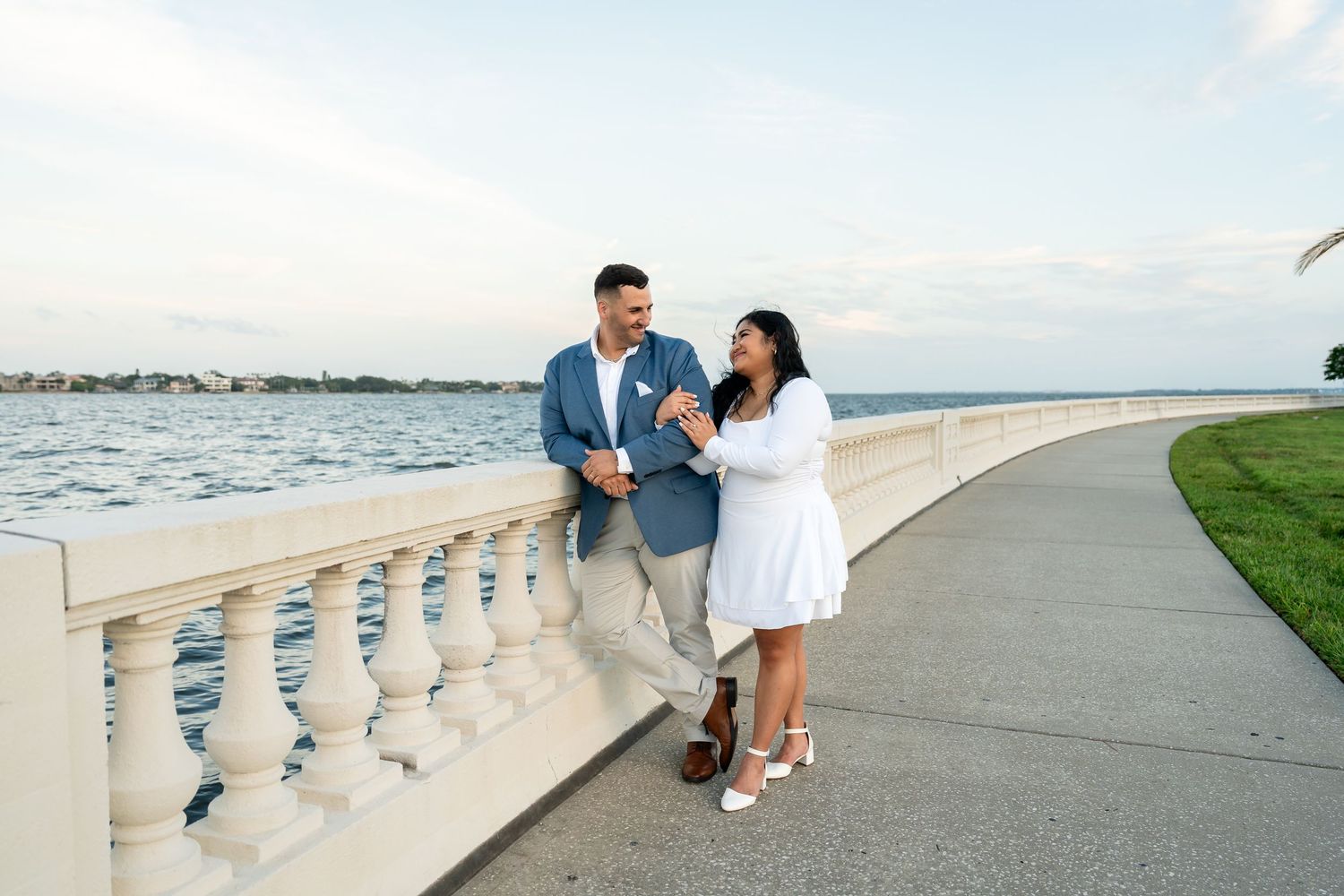 Couple walking along a waterfront promenade, smiling and holding hands, dressed in white and blue outfits at sunset.