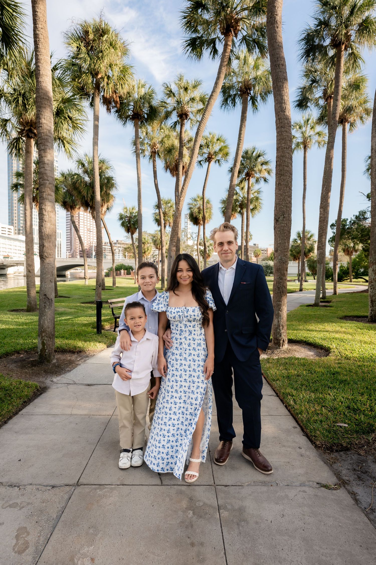 Family of four posing on a palm tree-lined path with blue sky and city buildings in background.