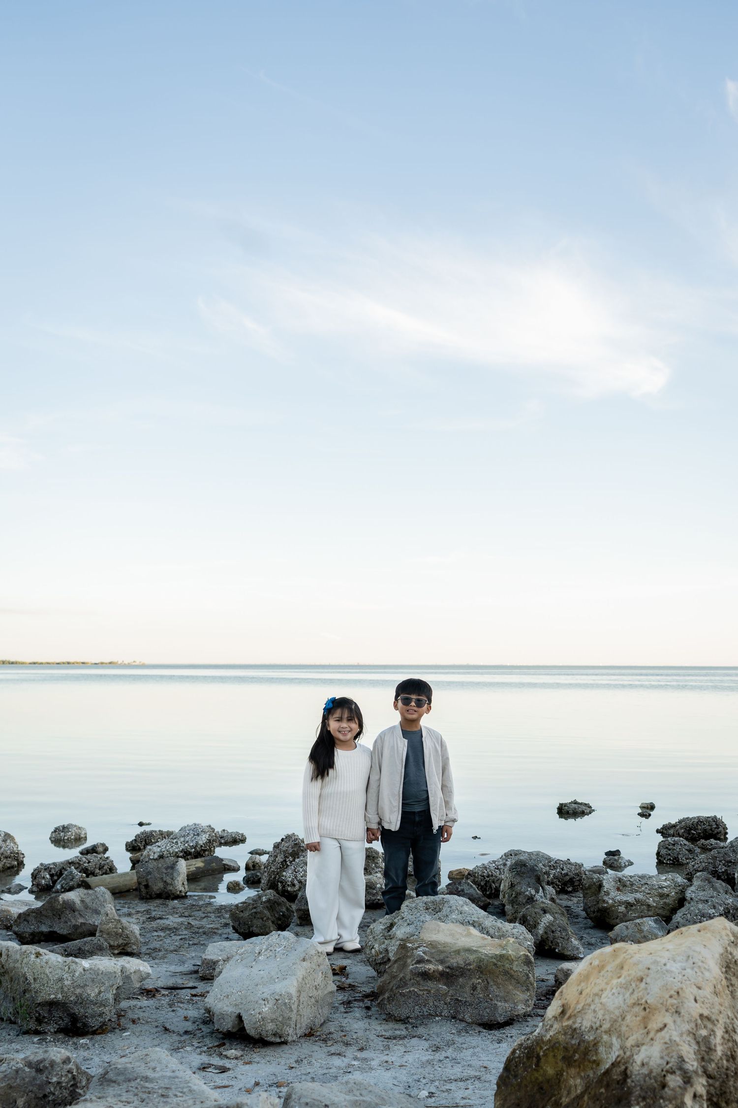 Two young children standing on rocky shoreline with calm ocean water and blue sky behind them.