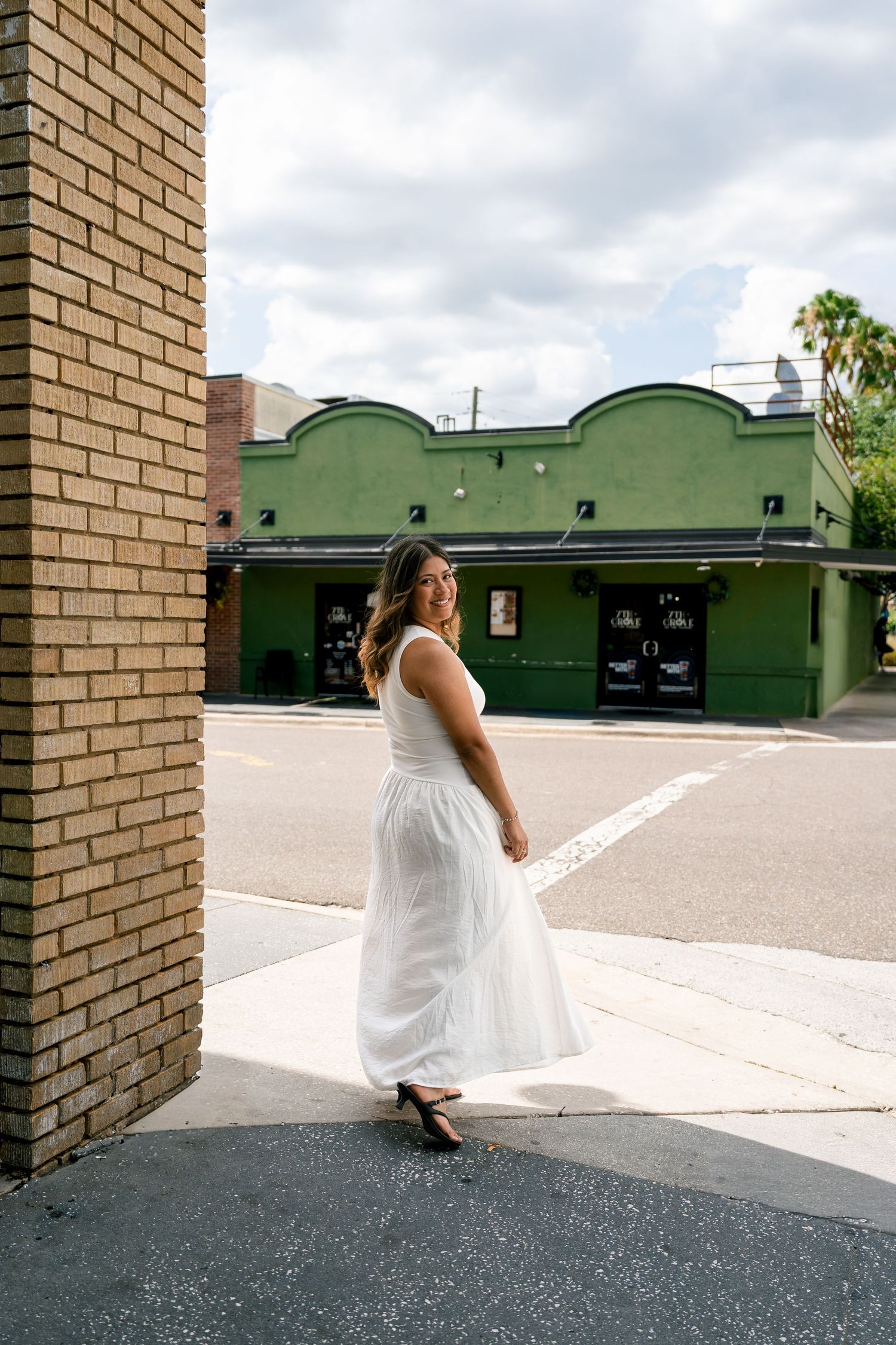Woman in white dress posing on urban sidewalk near brick building with green storefront backdrop.