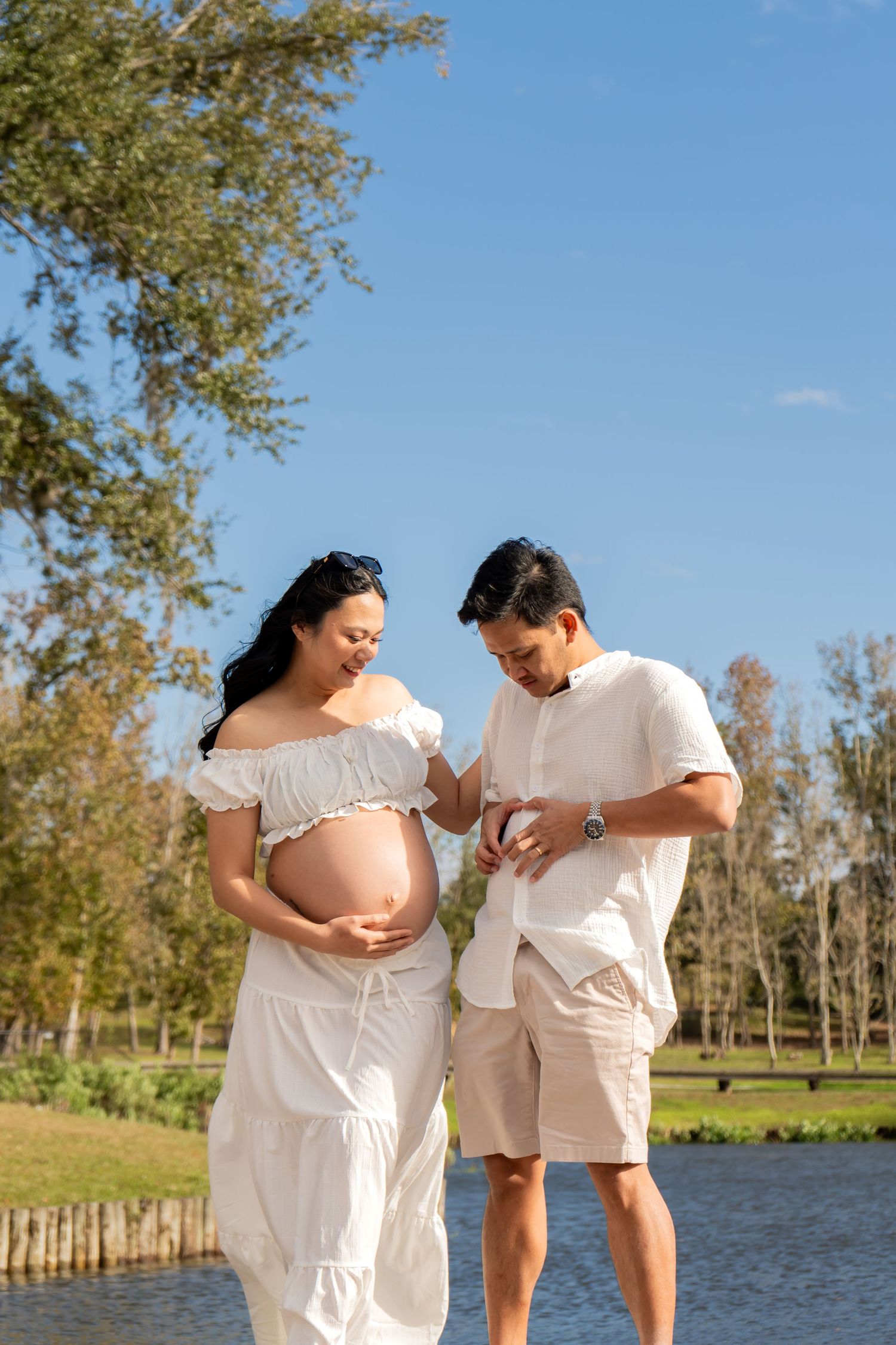 Couple expecting a baby standing by a lake, man touching pregnant womans belly on a sunny day.