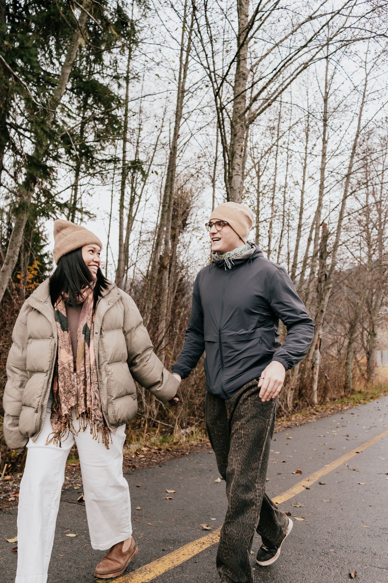 A couple holding hands and laughing while walking on a rainy path surrounded by bare autumn trees.