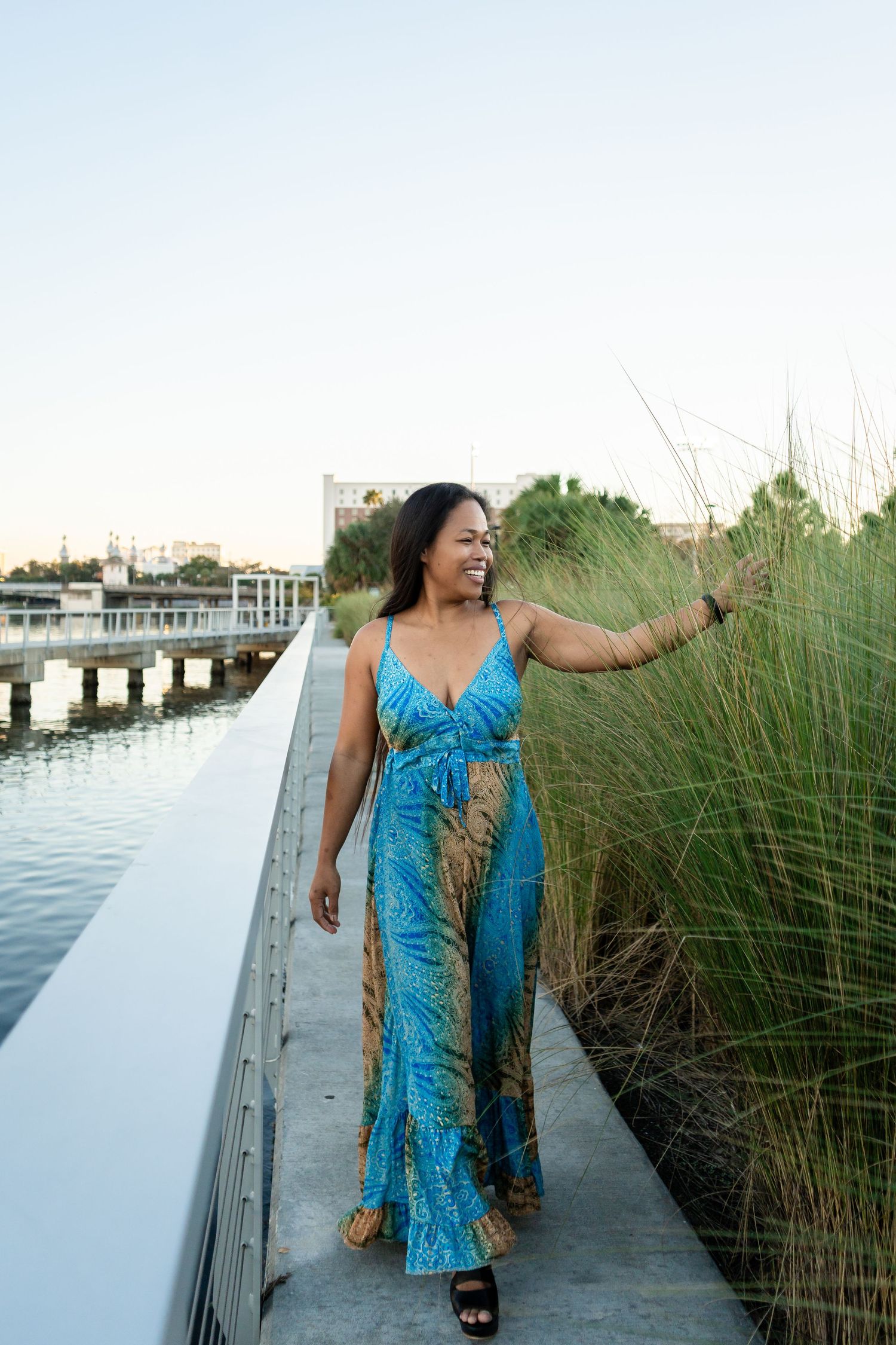 Woman in blue patterned jumpsuit walking along a waterfront path with tall ornamental grasses.