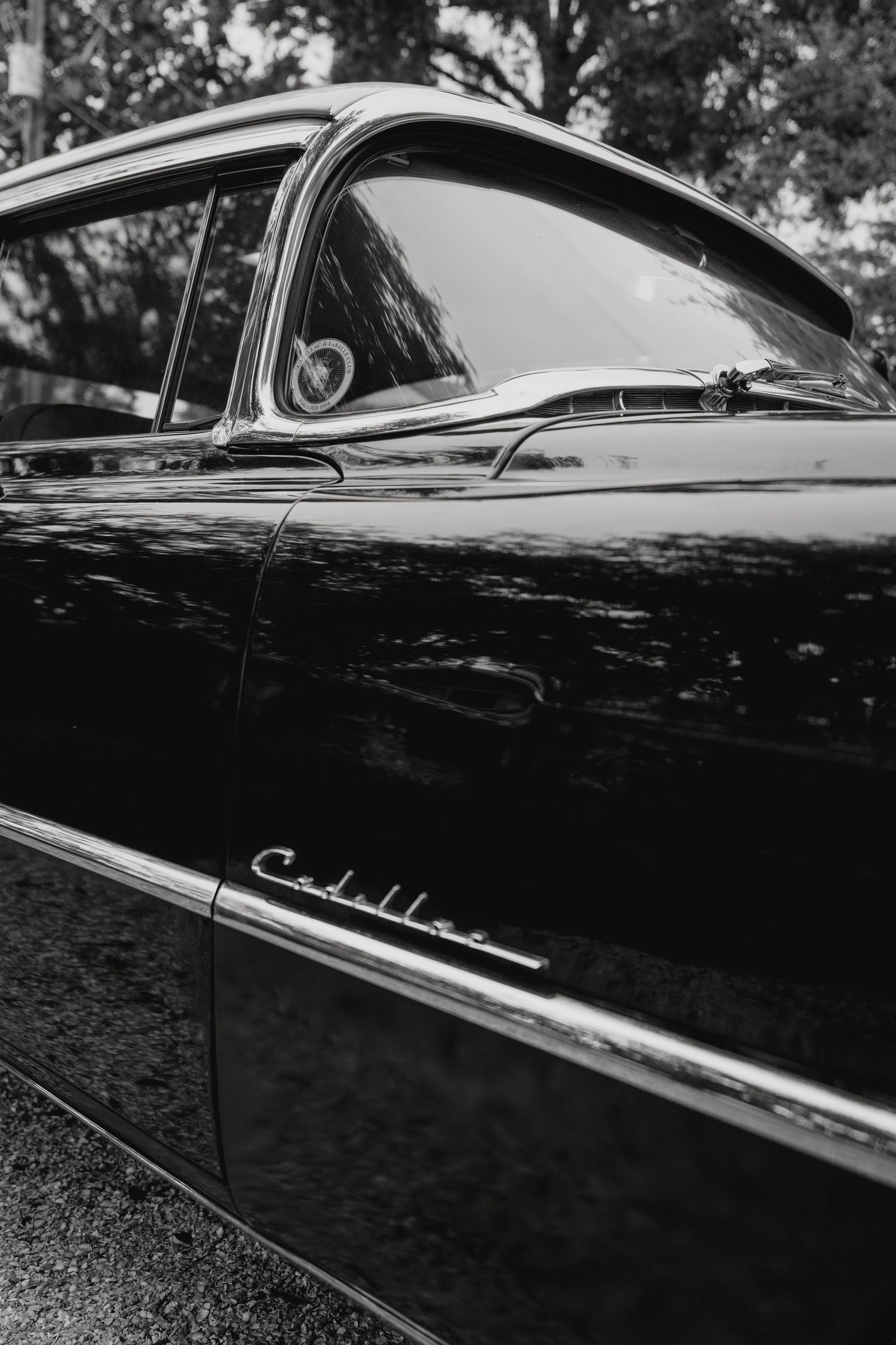 Black and white close-up of a vintage Cadillac car door with chrome emblem and reflective surface.