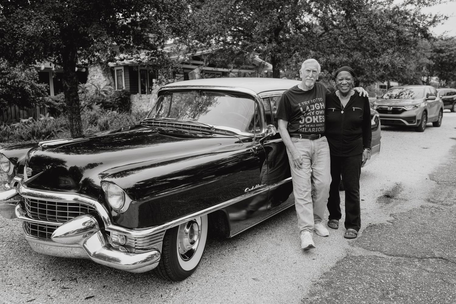 Two men posing beside a classic black 1955 Cadillac on a residential street.