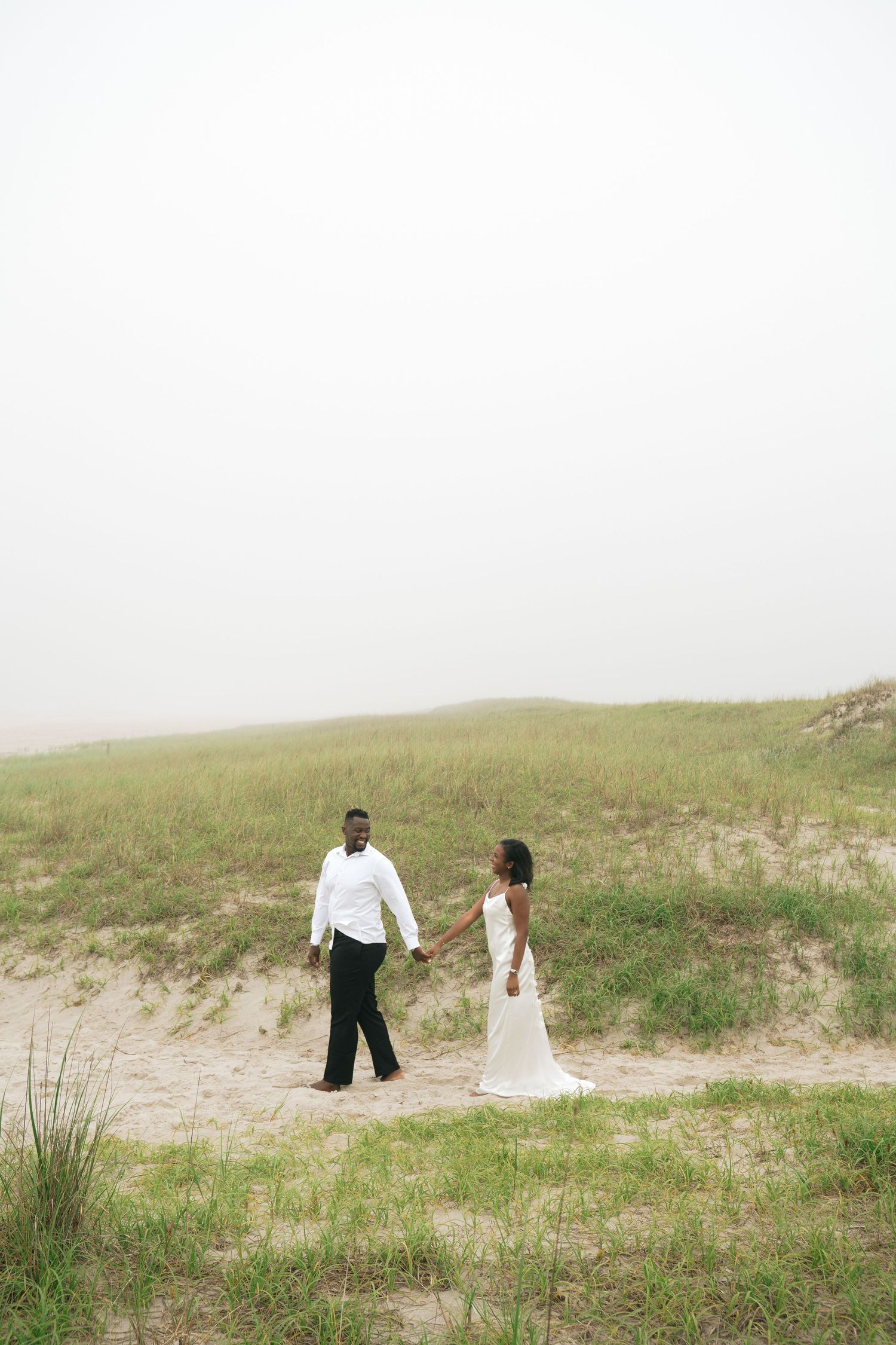 A couple in wedding attire holds hands on a sandy beach path surrounded by green dunes on a misty day.