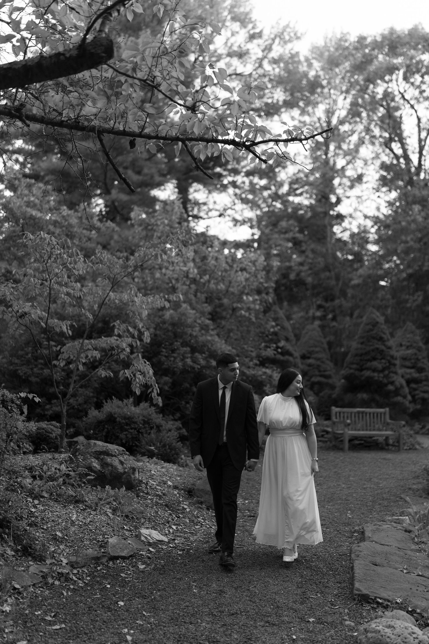 A couple walks together on a garden path surrounded by lush trees and foliage in black and white photography.