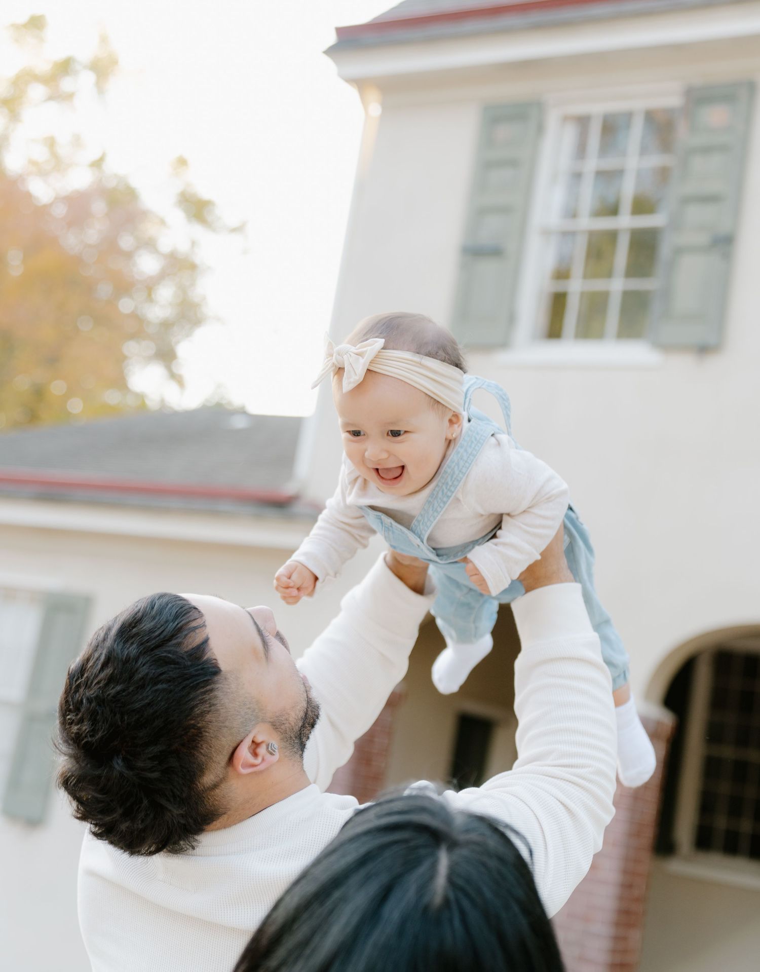 A parent lifts and plays with a baby wearing white against a white building exterior on a sunny day.