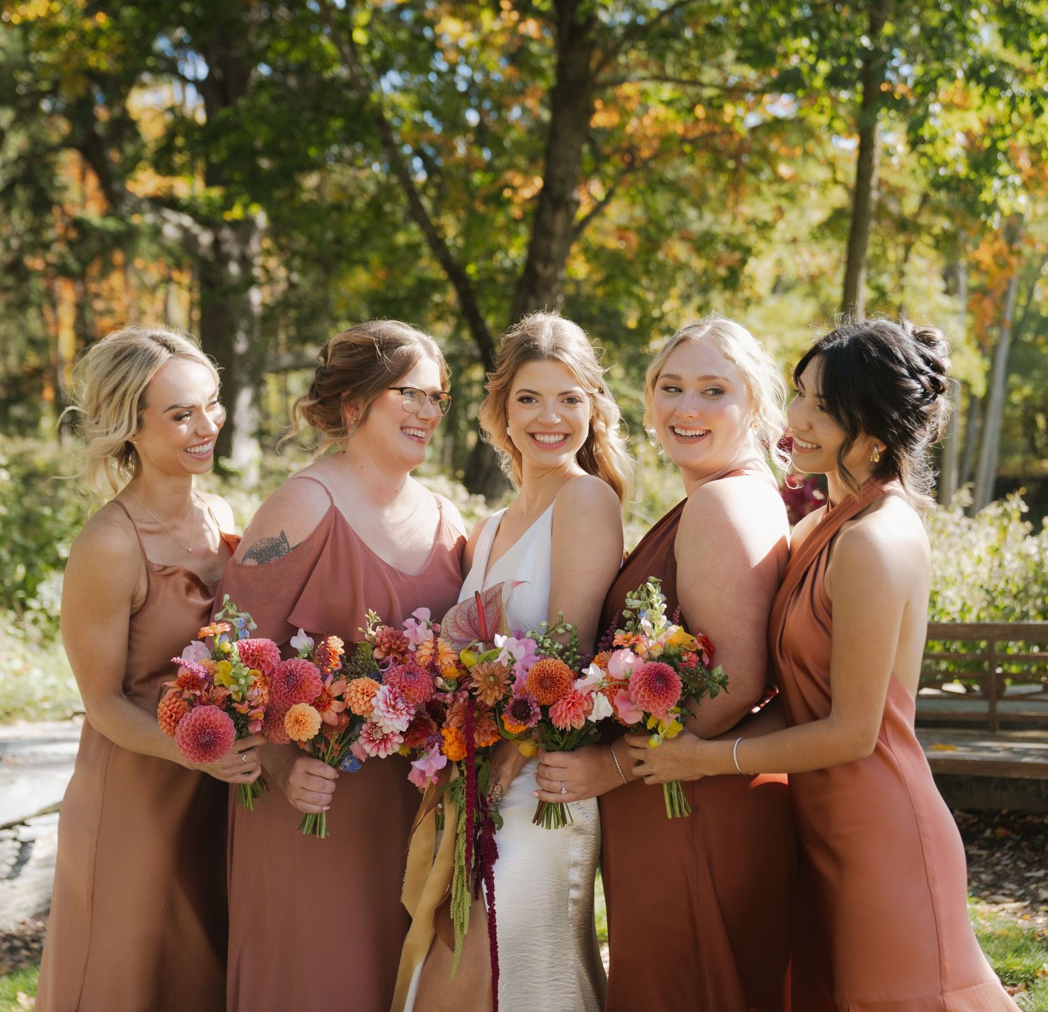 Bridesmaids in dusty rose dresses hold colorful bouquets while laughing together outdoors.