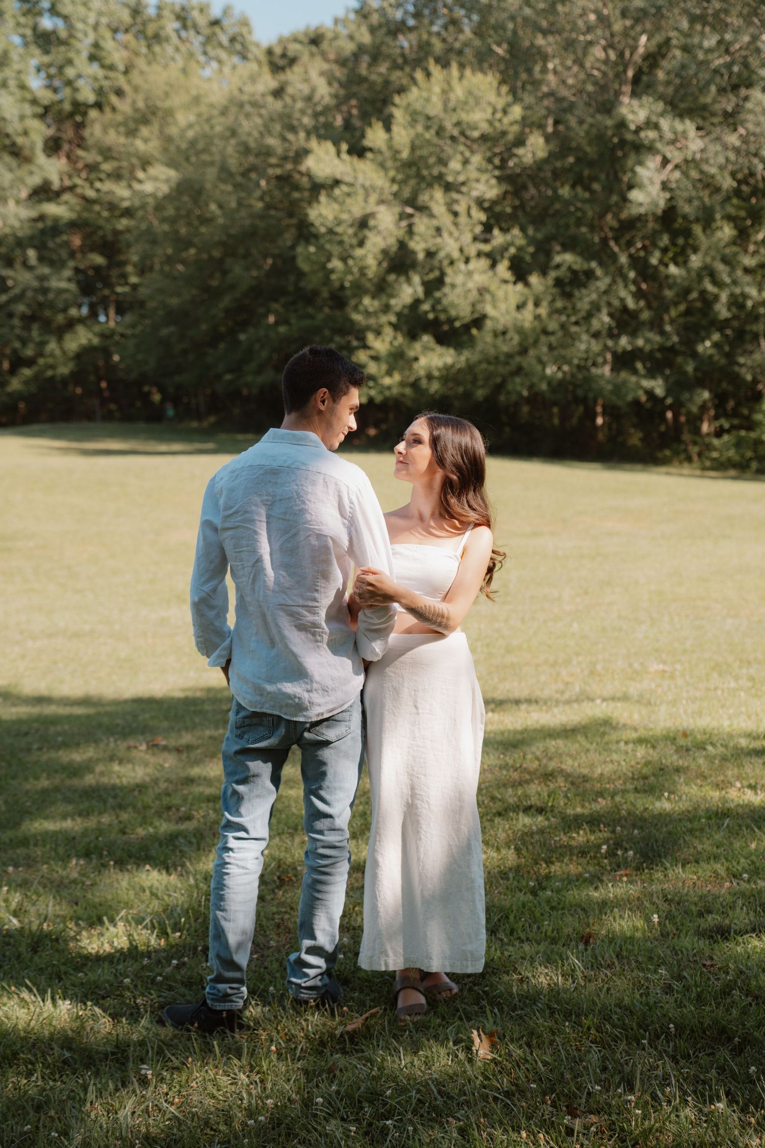 A romantic couple embraces in a sunny field surrounded by trees.