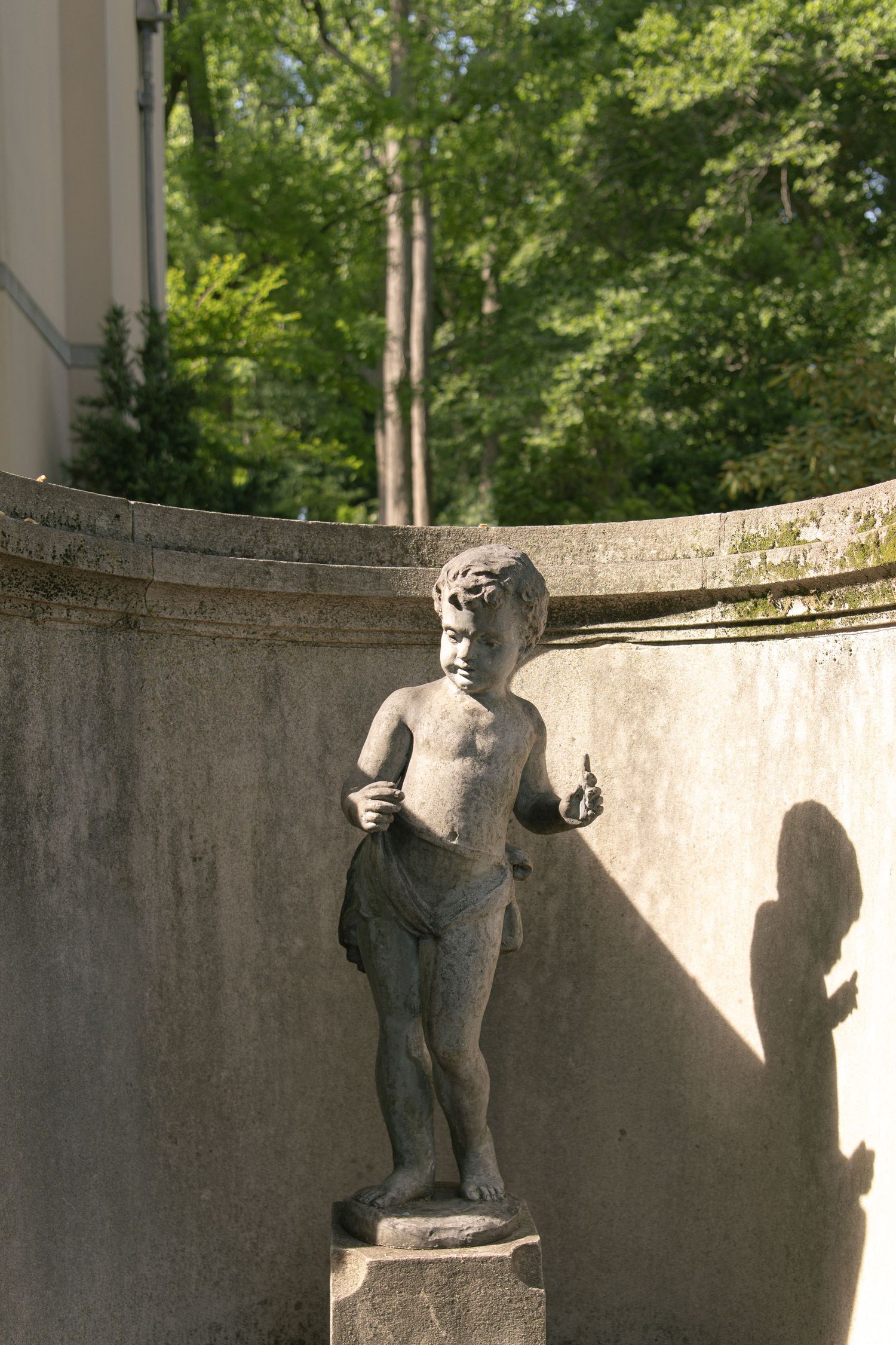 Stone cherub statue on pedestal against curved concrete wall with lush green trees in sunlit garden background.