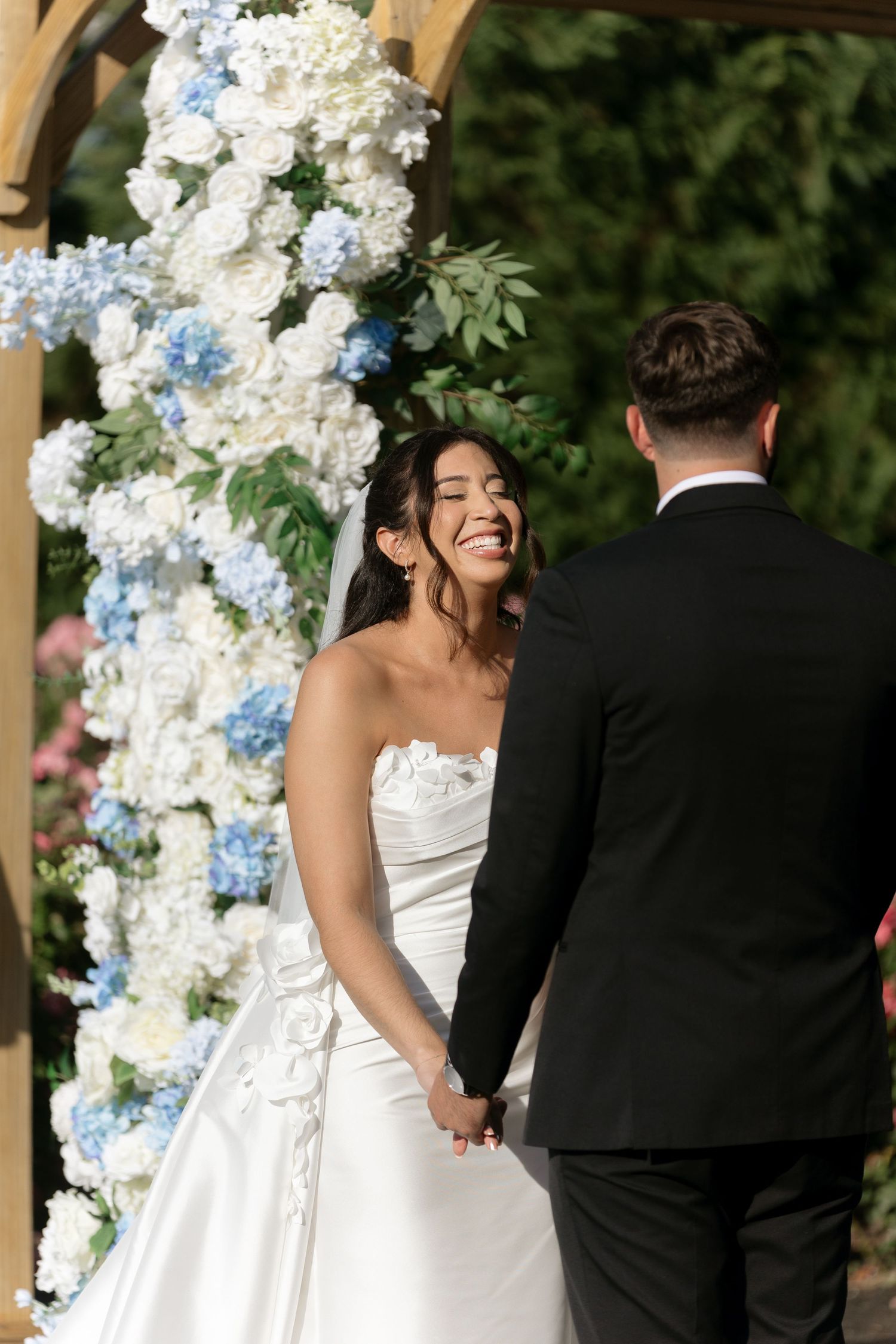 Joyful bride in white floral gown laughs holding grooms hands at blue and white floral altar.