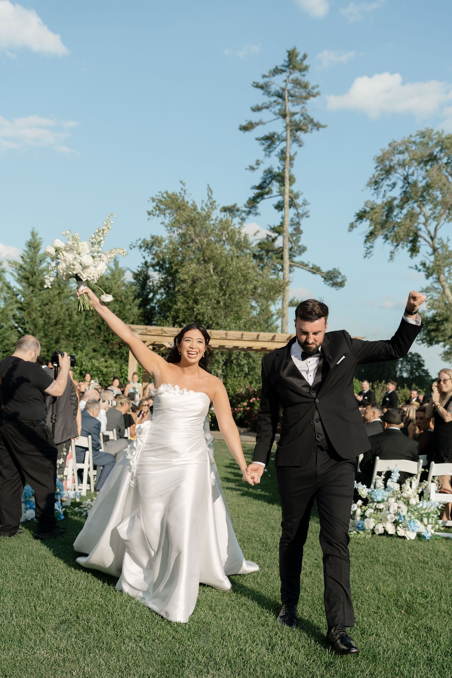 Joyful newlyweds celebrate their outdoor wedding ceremony, bride in white gown holding bouquet, groom in black tuxedo.