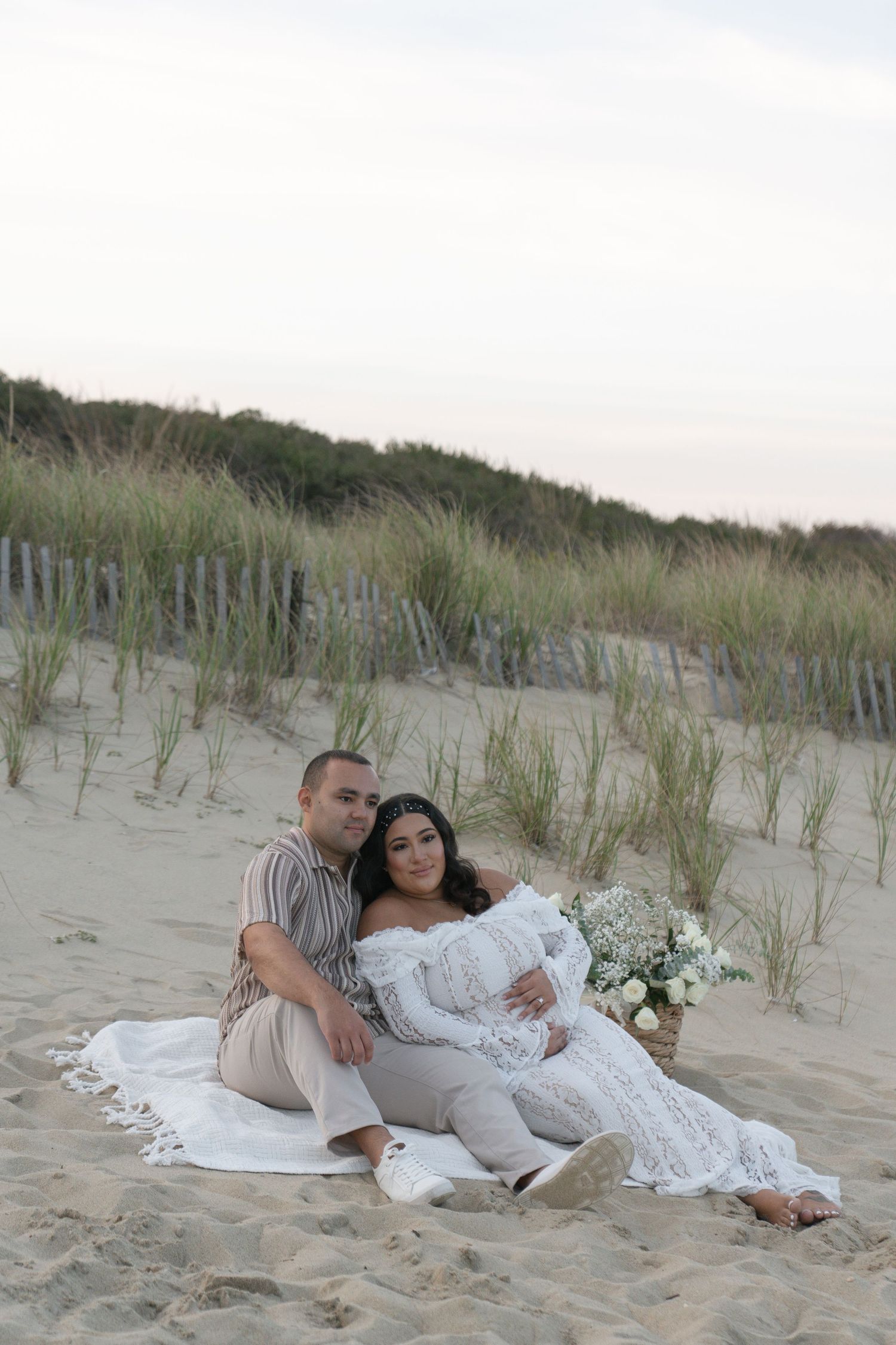 Couple sitting on beach sand dunes, woman in white lace maternity gown holding bouquet, man in striped shirt embracing her.