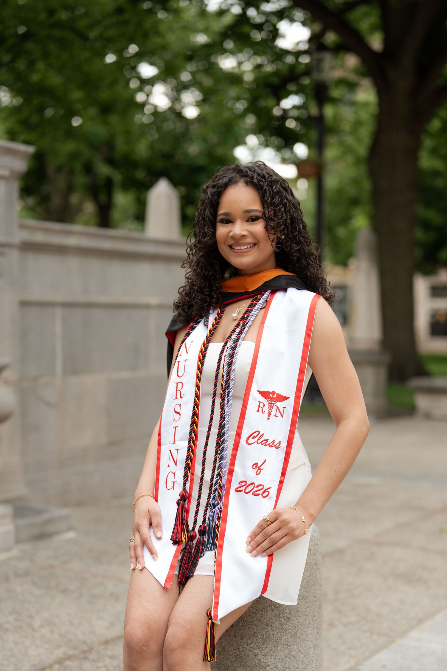 Nursing graduate smiling in white dress with RN Class of 2026 sash and colorful honor cords outdoors.