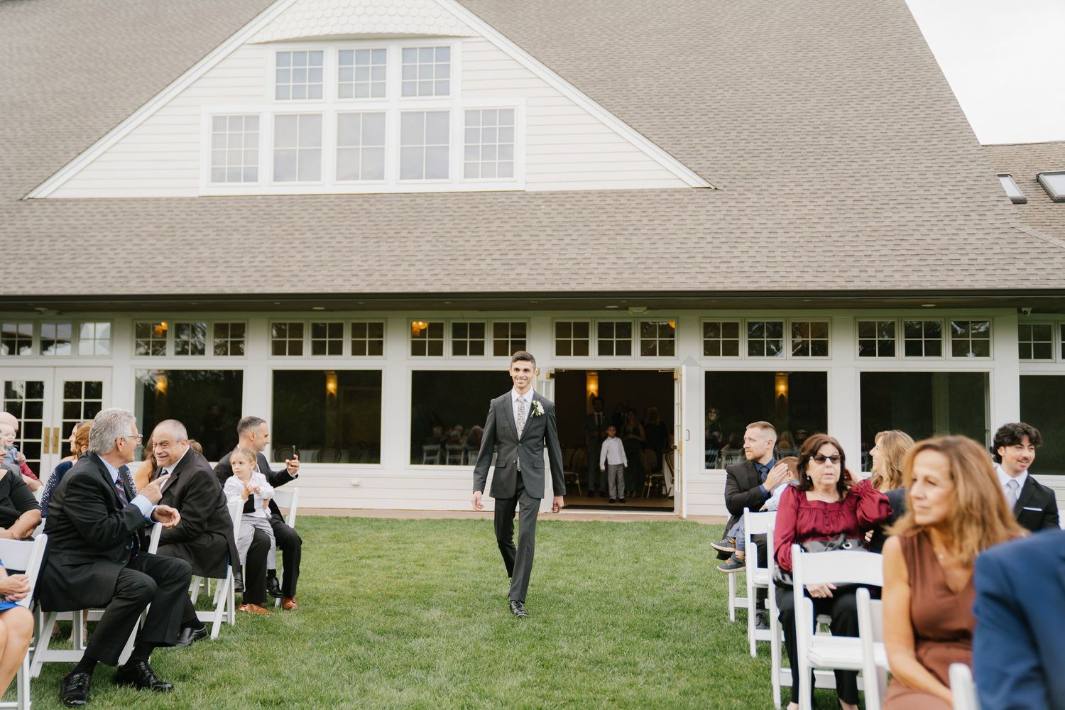 Wedding guests gather on a green lawn outside a white building with dormer windows during an outdoor celebration.