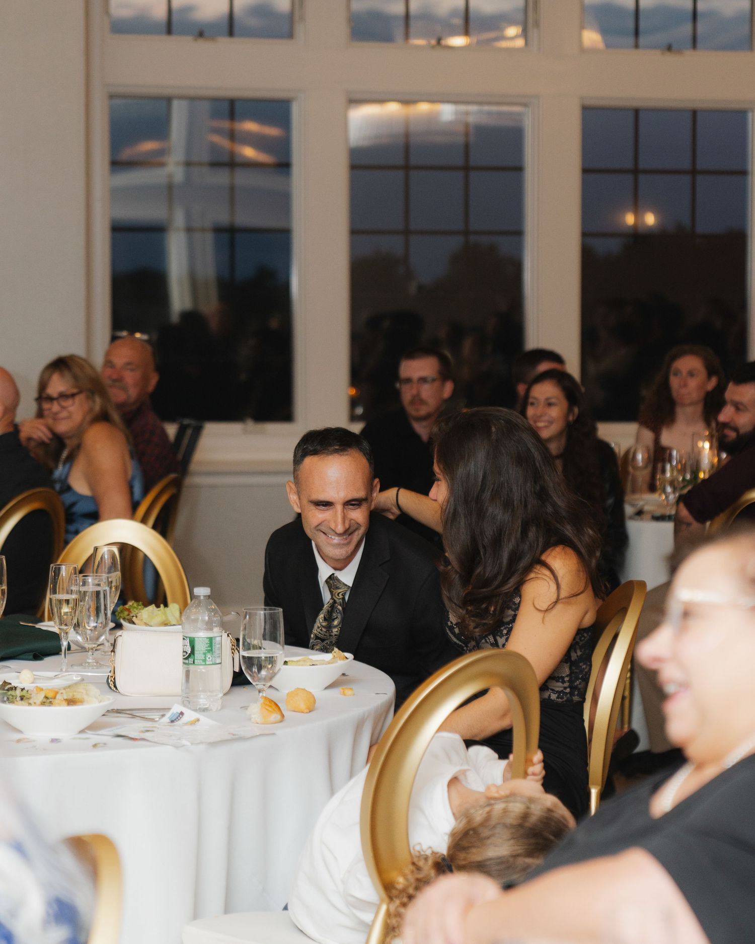 Guests seated at round tables with white tablecloths during an elegant evening reception with large windows in background.