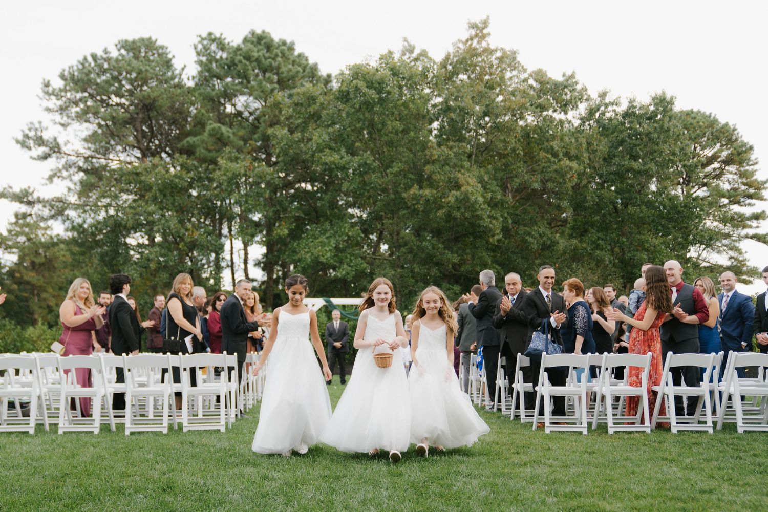 Panoramic view of an outdoor wedding ceremony with white chairs and guests gathered under trees.