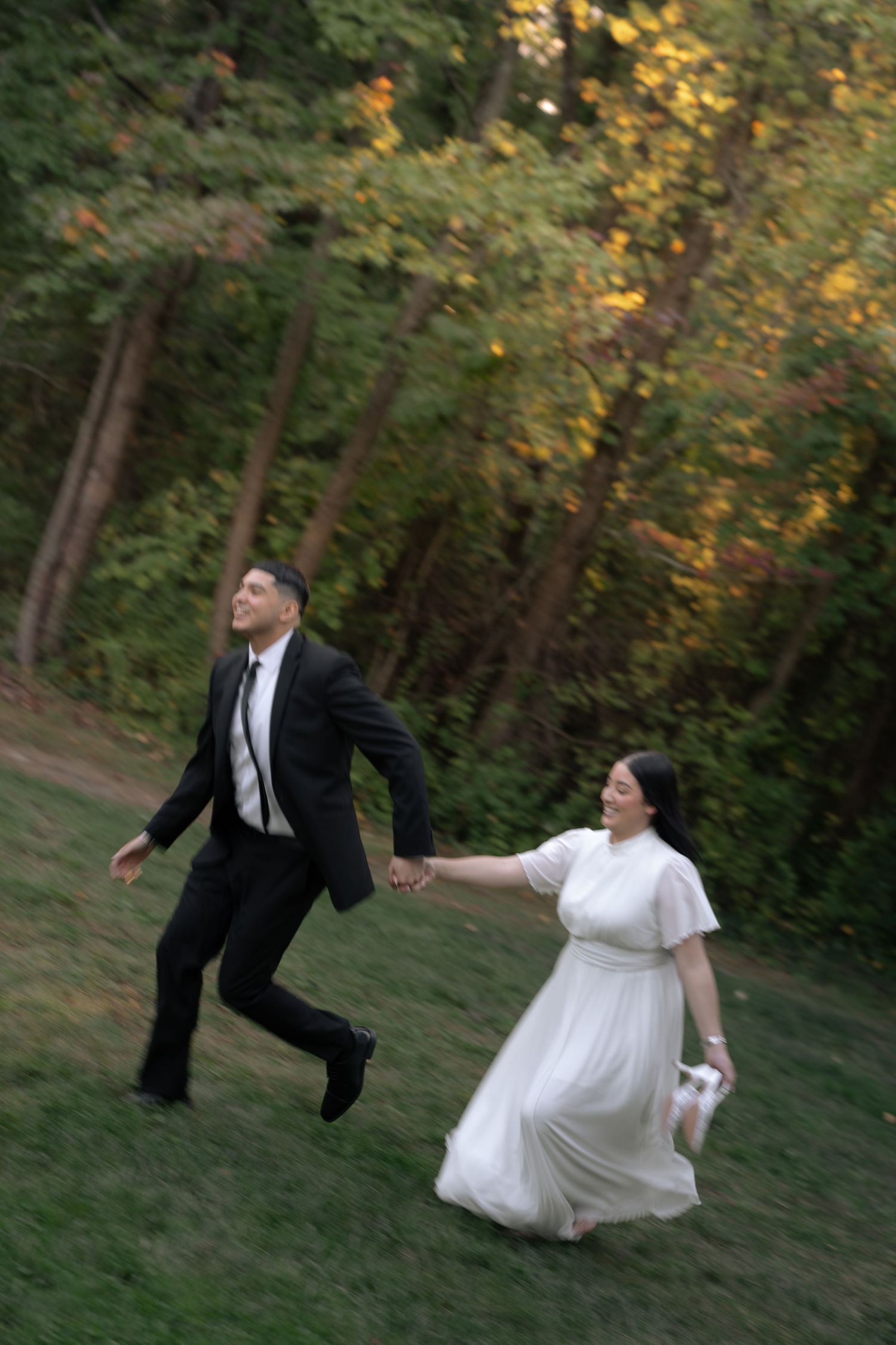 A couple in wedding attire playfully runs through a wooded outdoor setting at sunset.