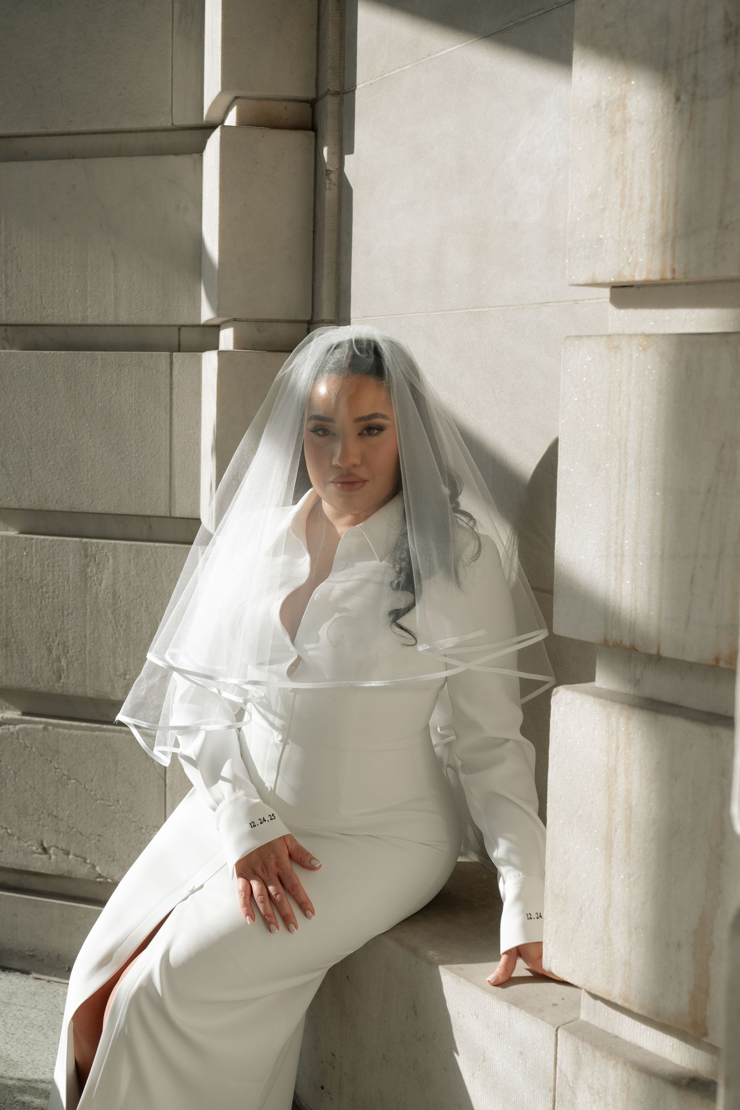 A bride in a modern white dress and veil poses against a sunlit architectural backdrop.
