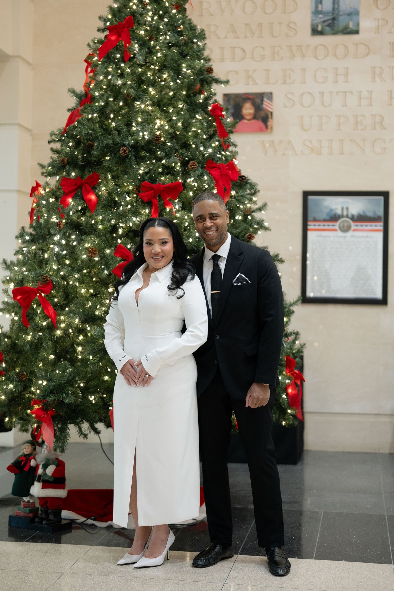 A couple poses in front of a decorated Christmas tree with red bows and twinkling lights in an elegant setting.