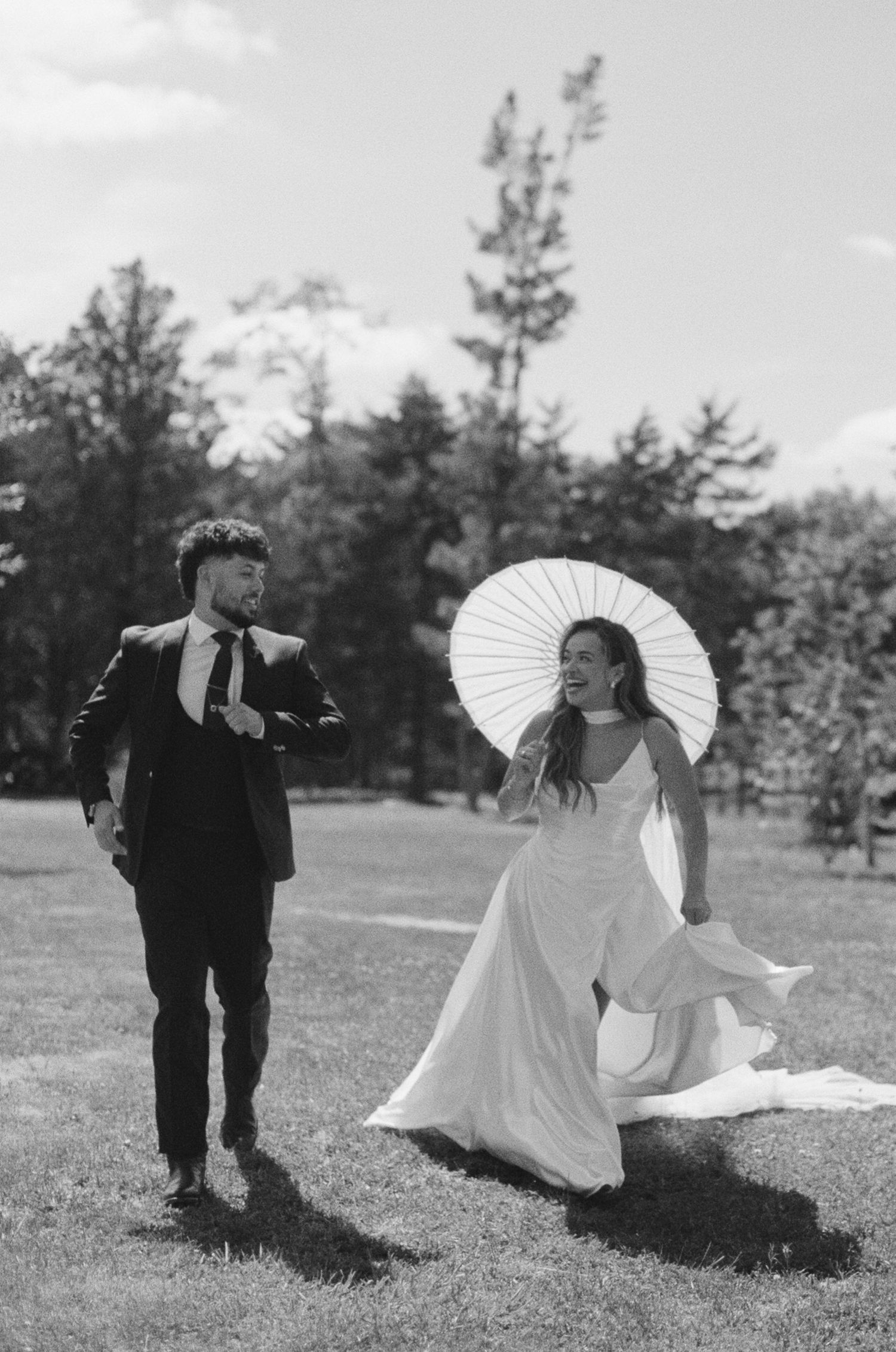 Black and white photo of a smiling bride holding a parasol in a flowing gown walking with a groom in a suit outdoors.