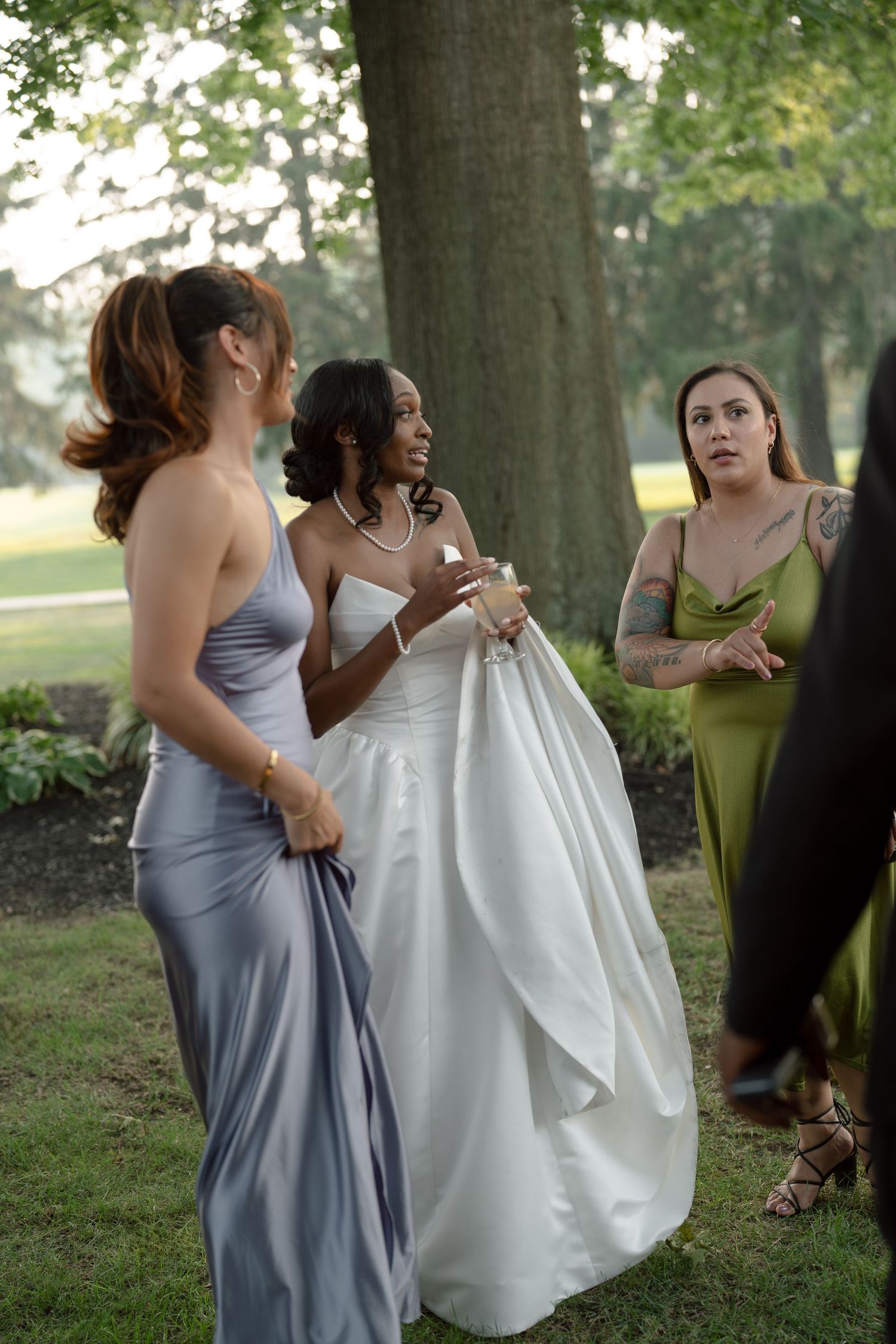 Bride in white ball gown chats with bridesmaids in silver and green dresses outdoors.