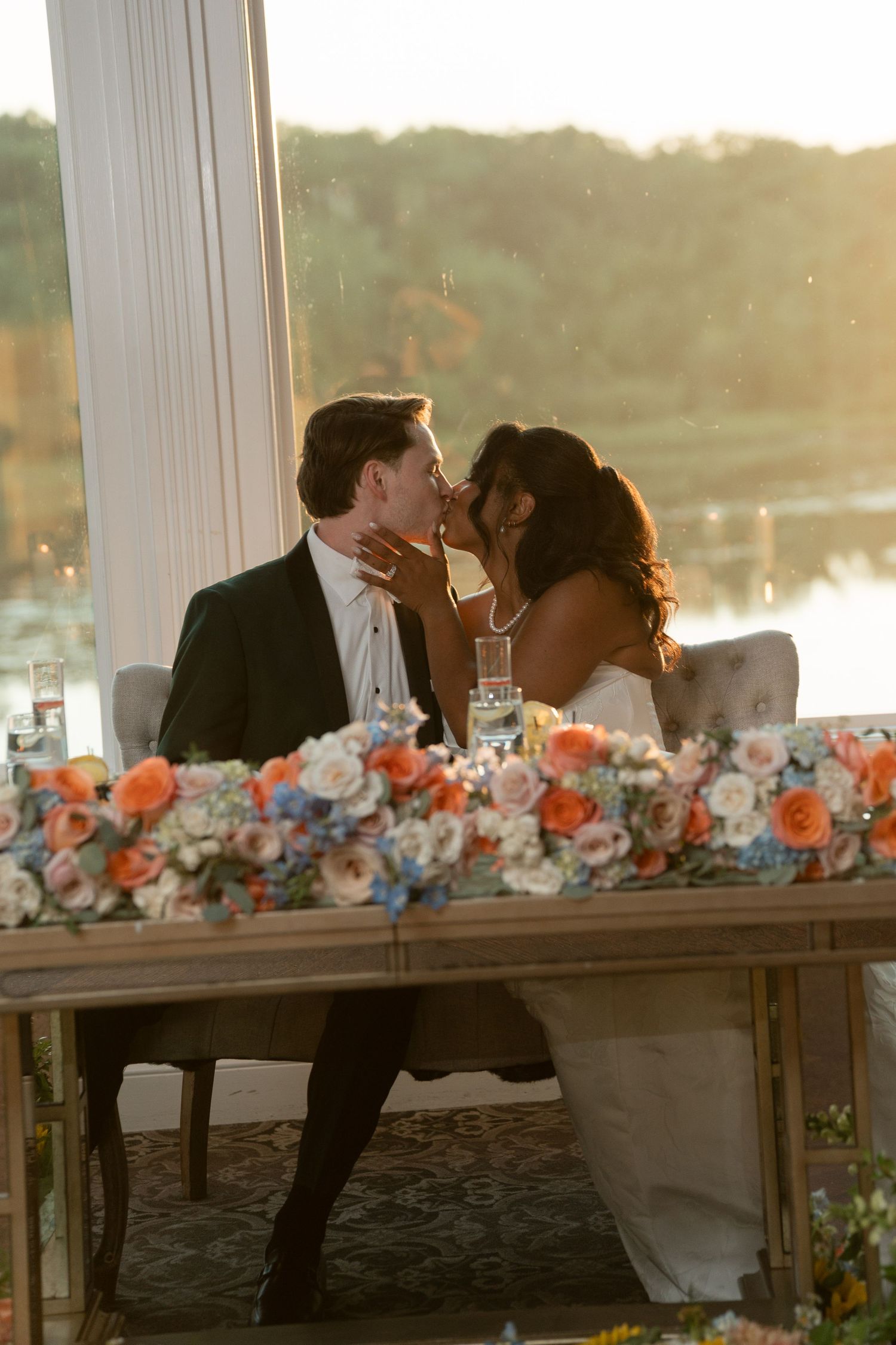 Newlyweds share a romantic kiss at their sweetheart table adorned with coral and blue floral arrangements at sunset.