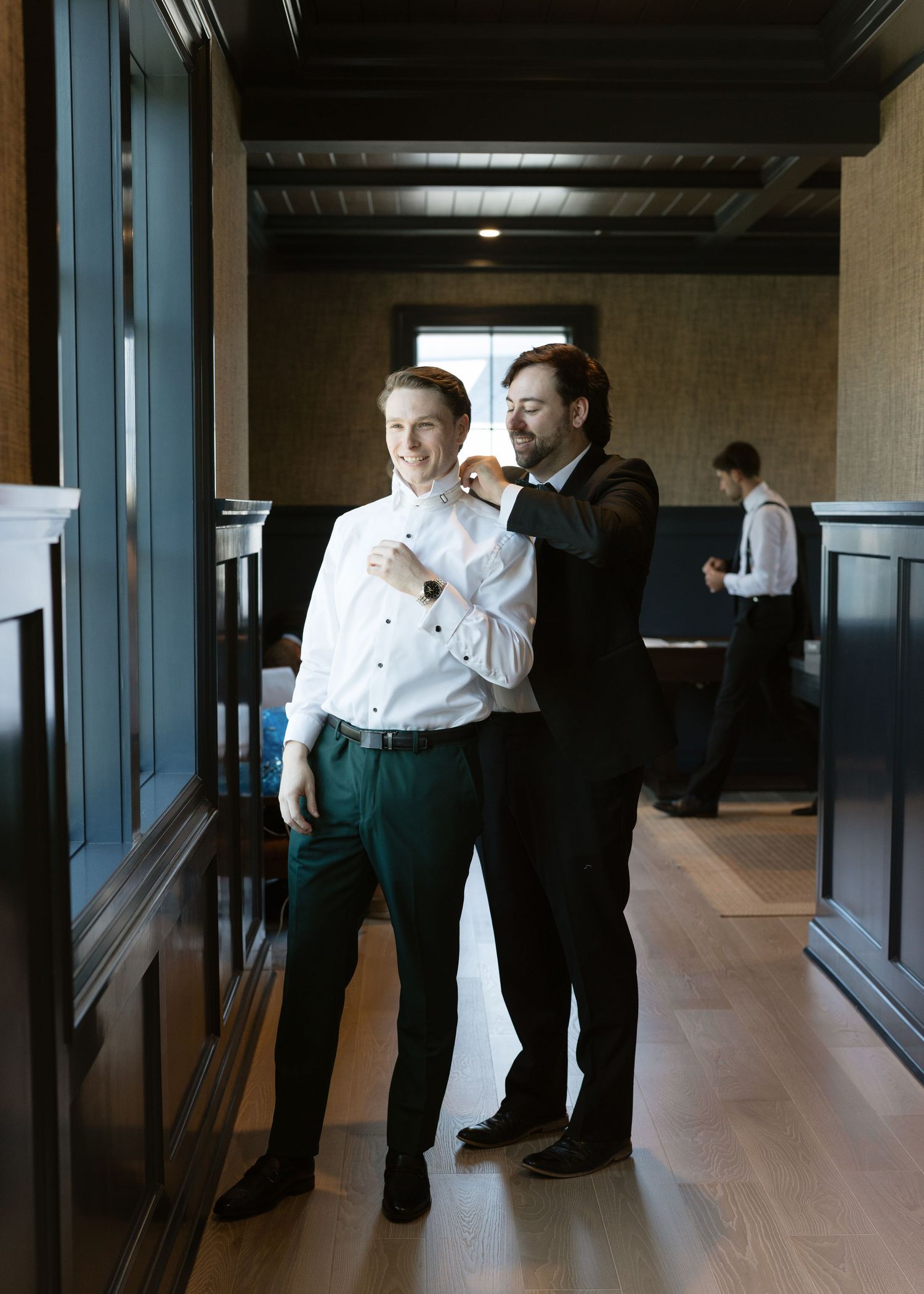 Groomsman helps groom adjust his collar while getting ready in an elegant dark wood-paneled room.
