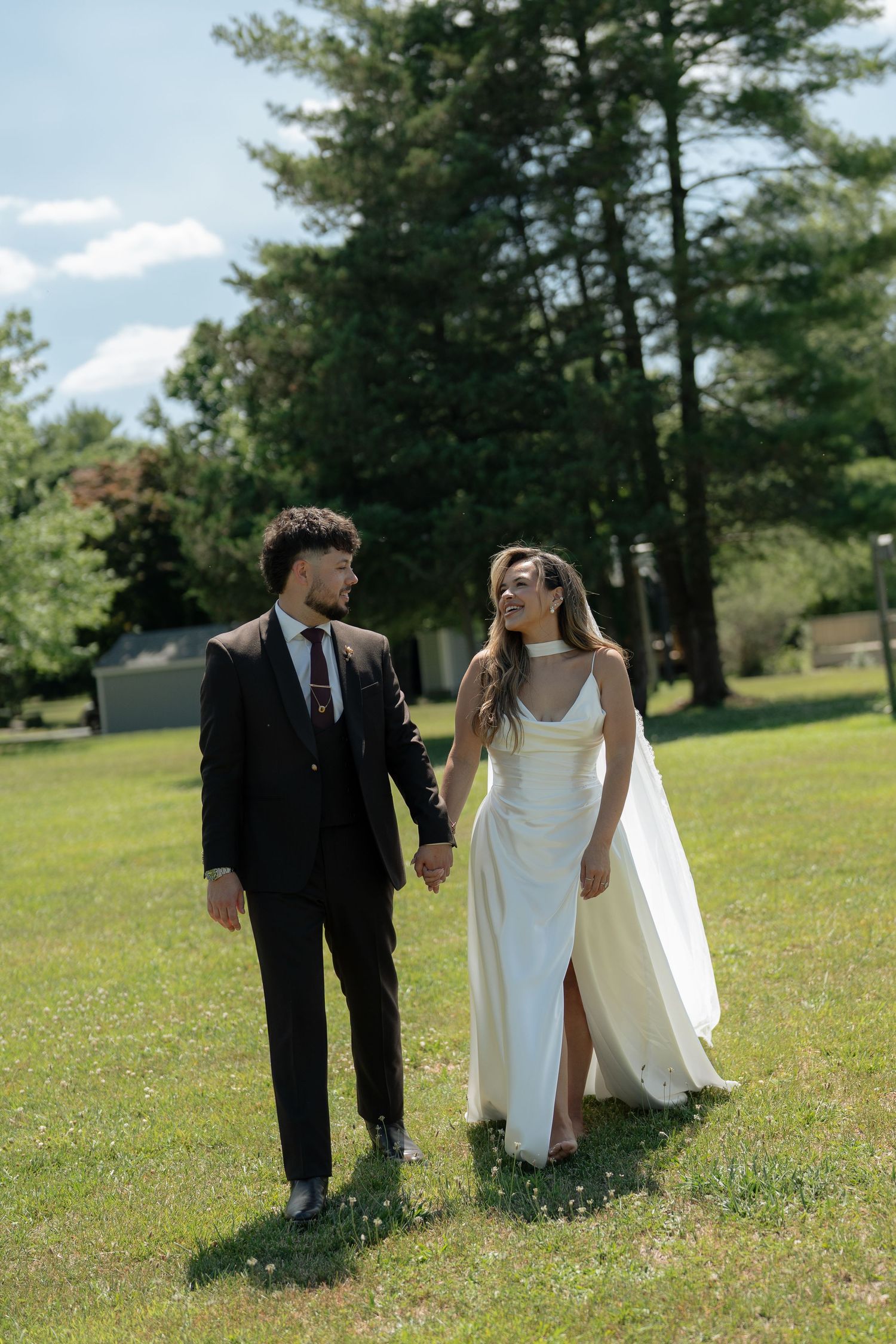 Bride and groom walk hand-in-hand across green lawn, smiling at each other on their wedding day.