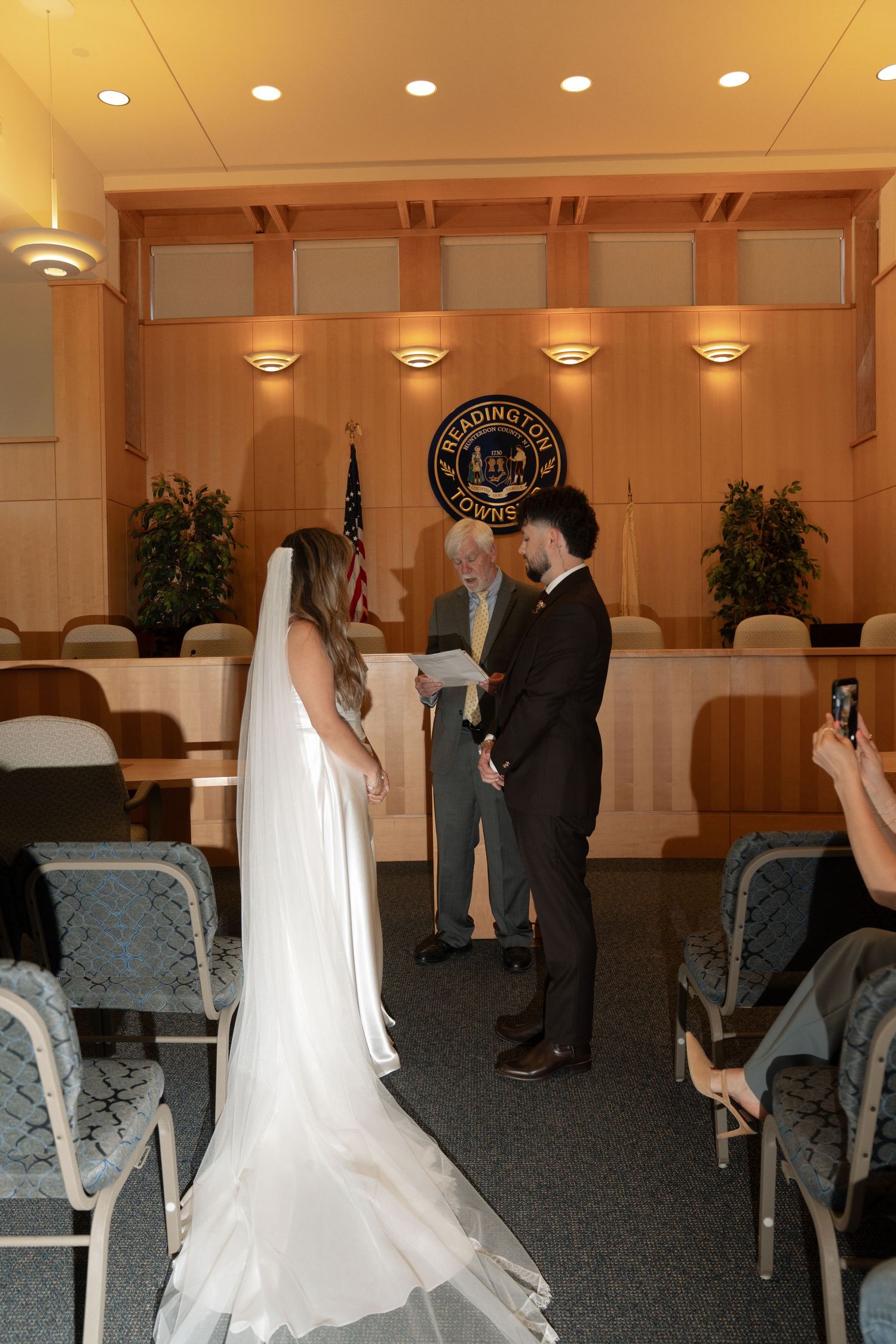 Bride and groom exchange vows before an officiant in an elegant wood-paneled ceremony room with warm lighting.