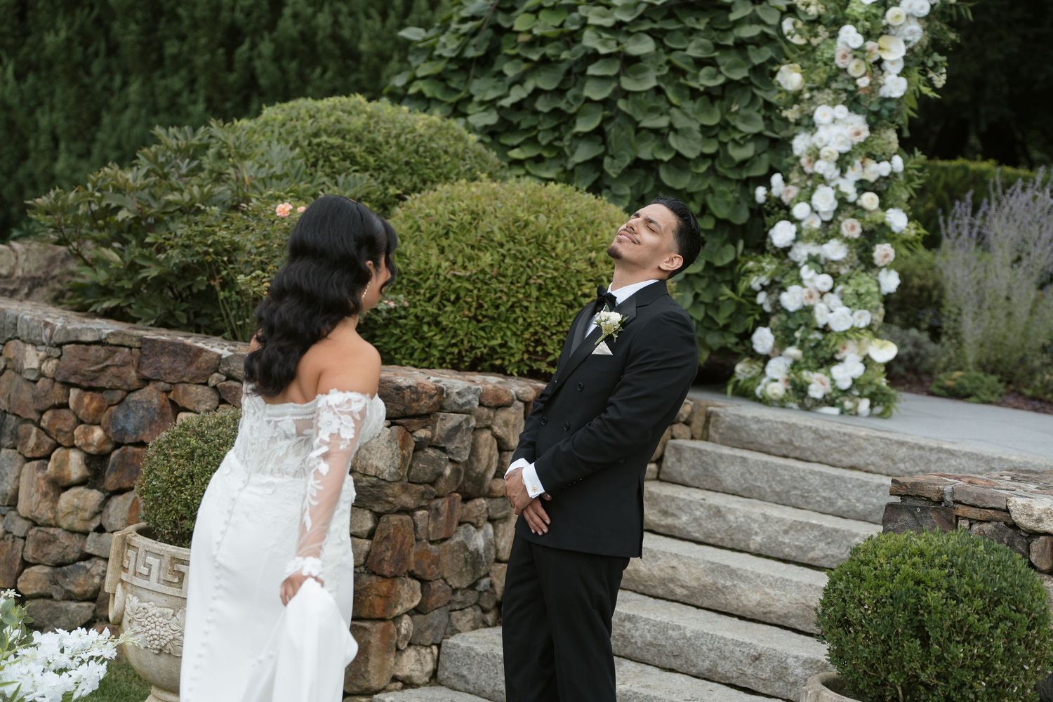 Groom in black tuxedo reacts emotionally during first look with bride in lace wedding gown in garden.