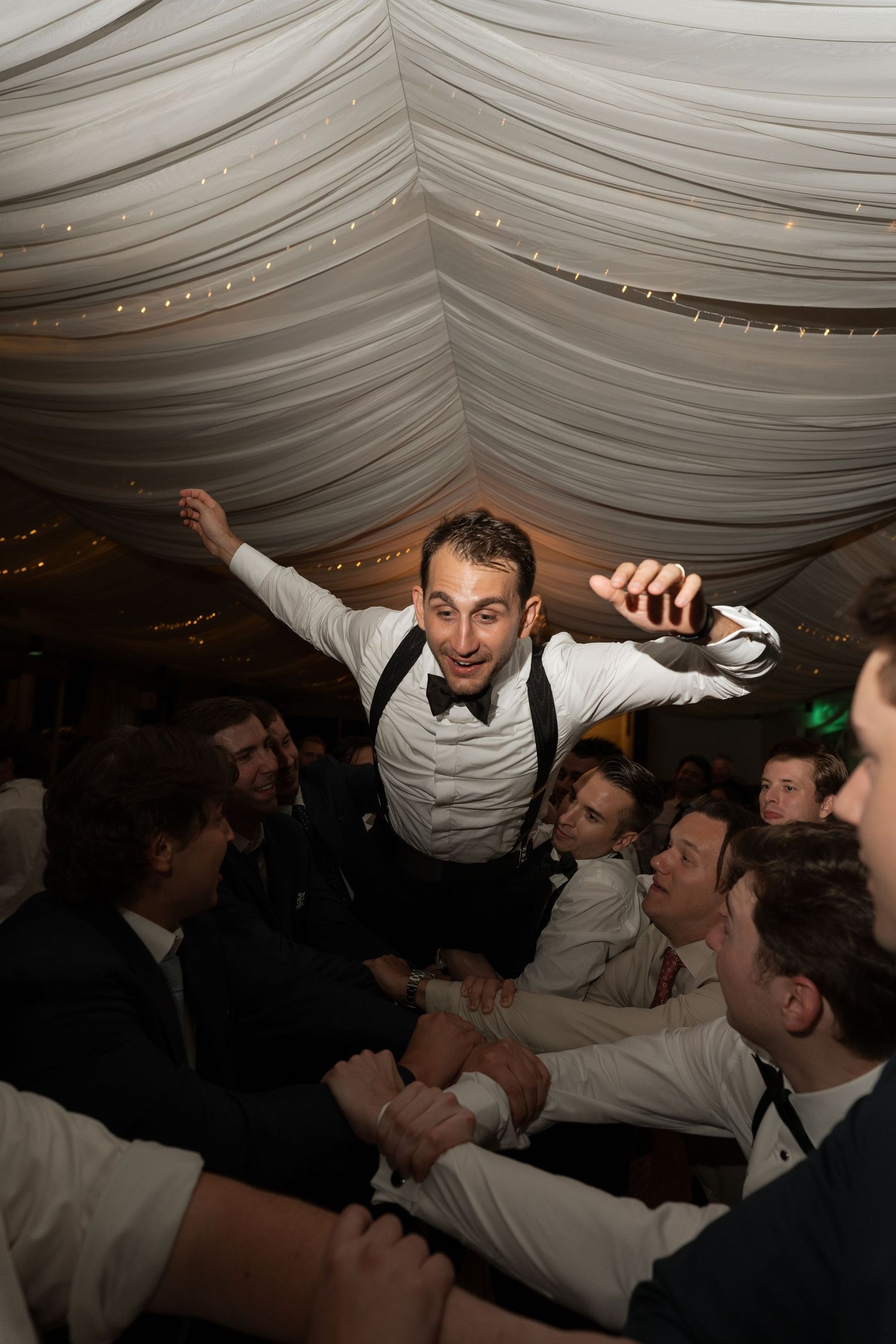 Groom is lifted by groomsmen in the air during energetic wedding reception dance celebration.