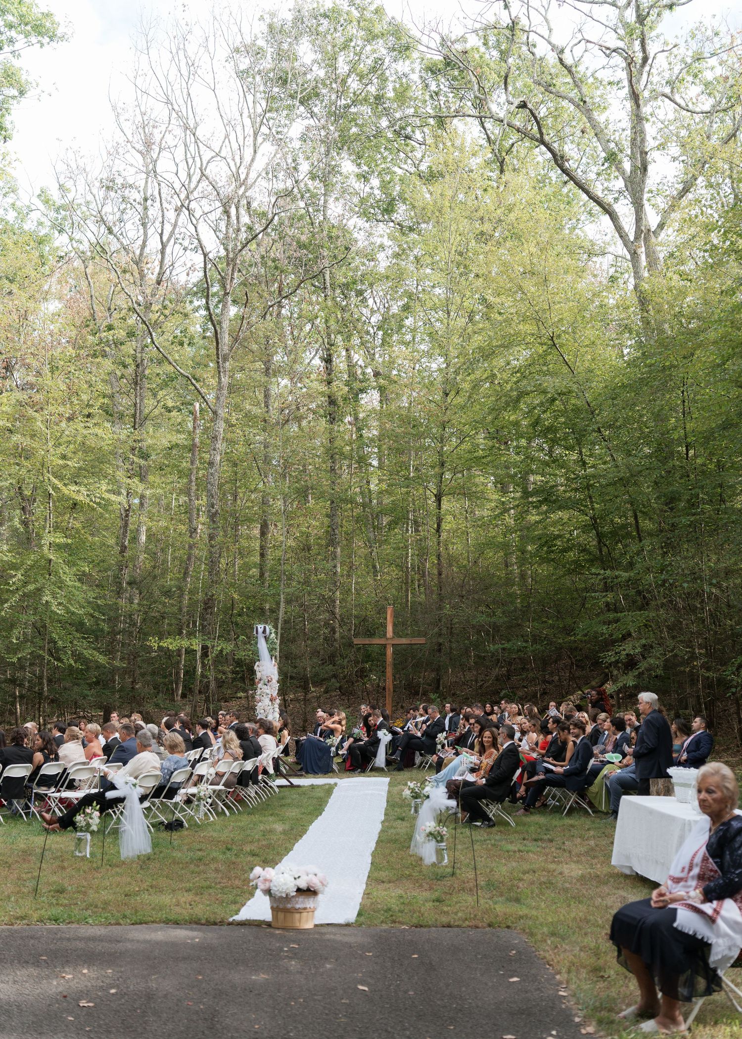 Outdoor wedding ceremony in a forest clearing with white chairs, a wooden cross, and a white aisle runner.