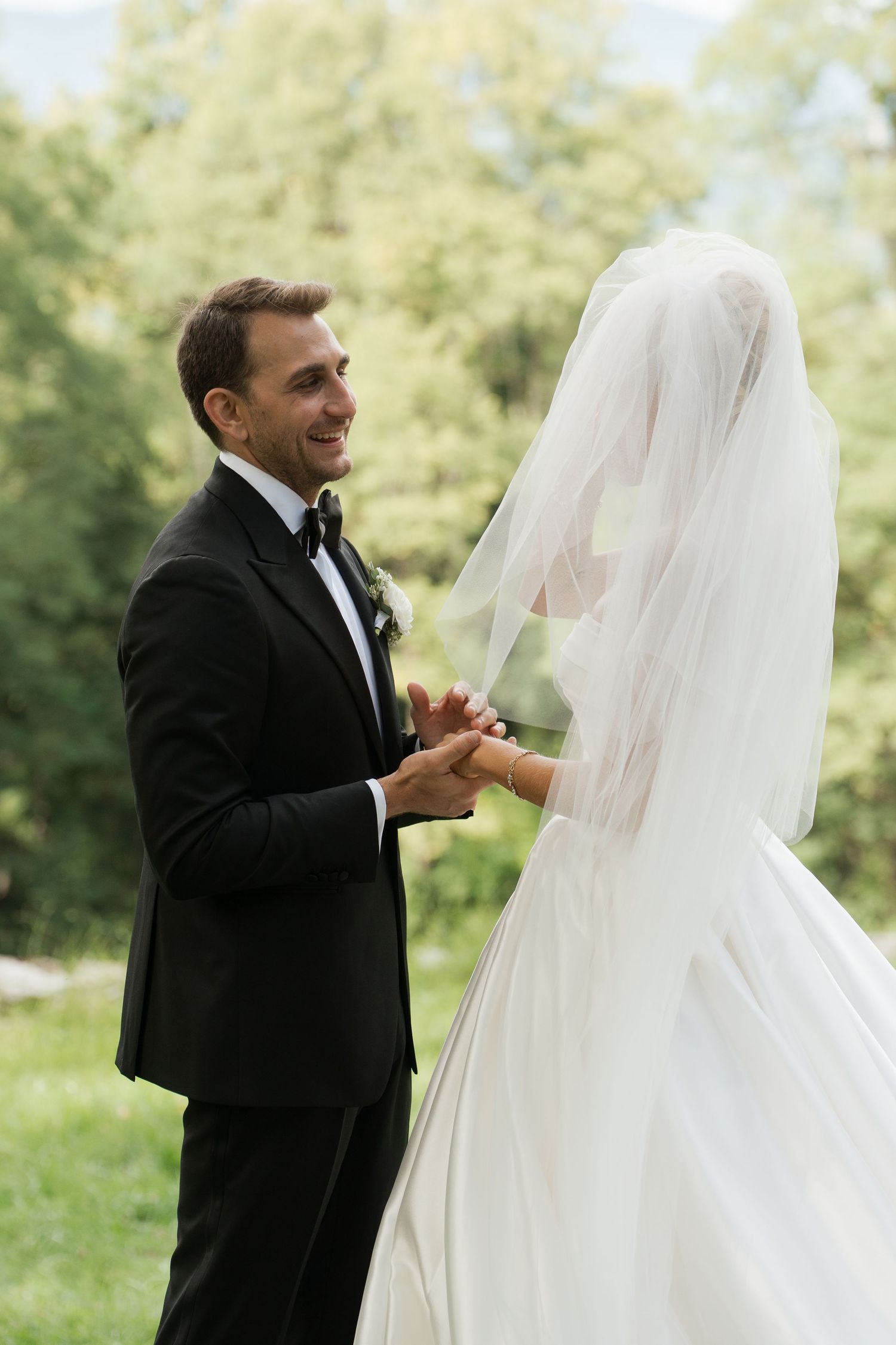 Smiling groom in black tuxedo holds hands with bride in white wedding gown during outdoor first look.