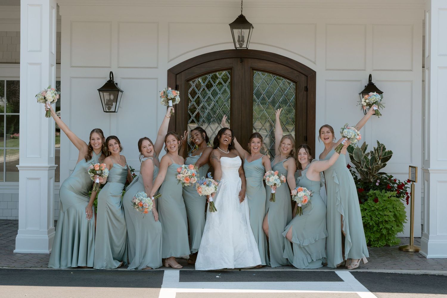 Bride in white gown poses with bridesmaids in sage green dresses holding bouquets outside elegant white venue.