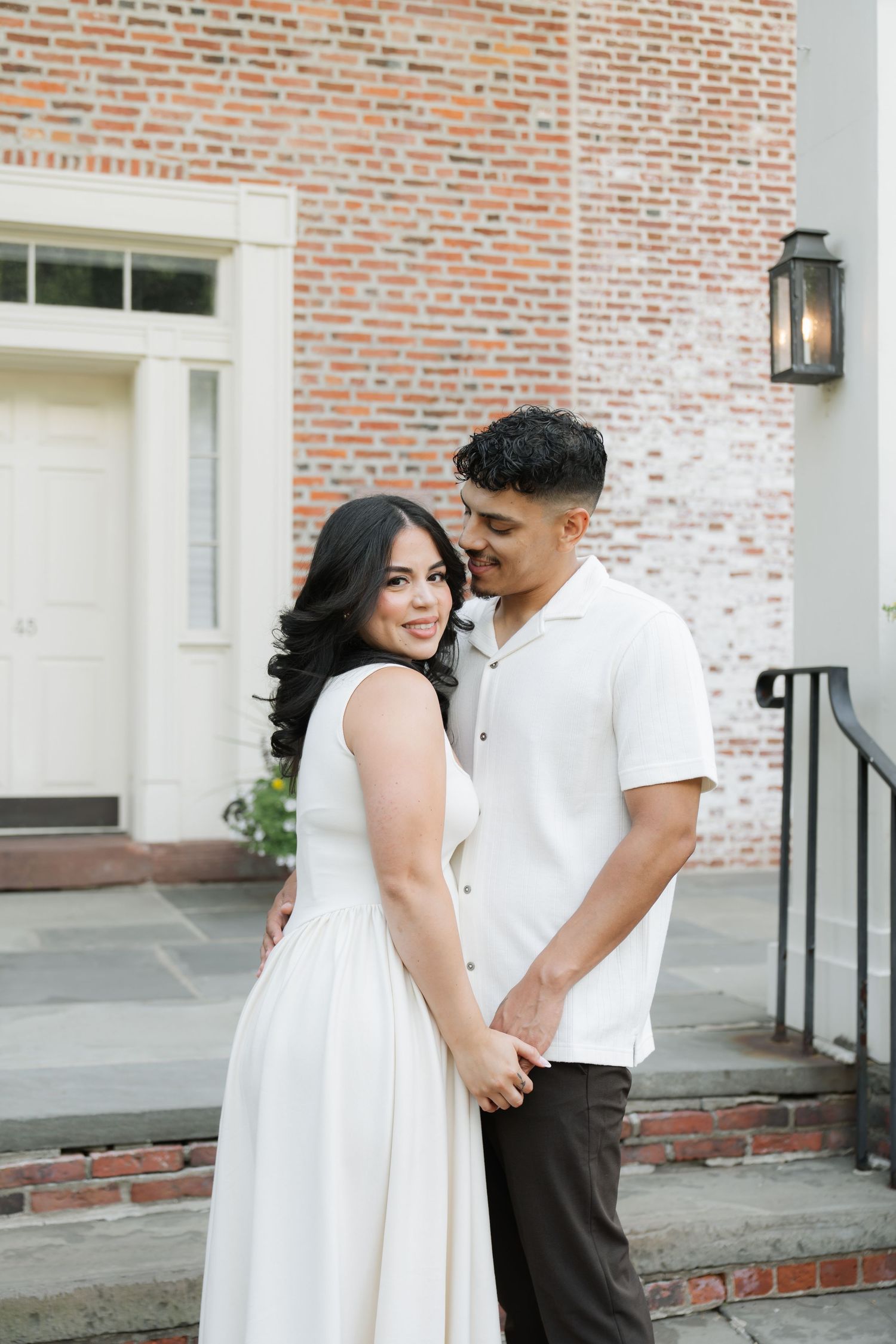 A couple in coordinated white outfits share an intimate moment in front of a brick building with a lantern.