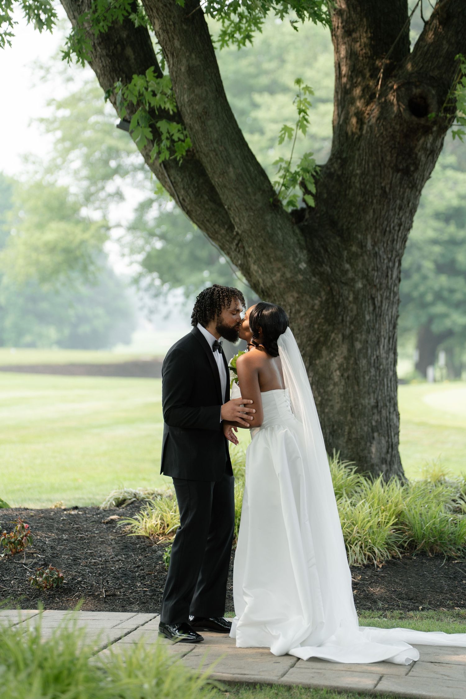 A bride and groom share a romantic kiss under a large tree on their wedding day outdoors.