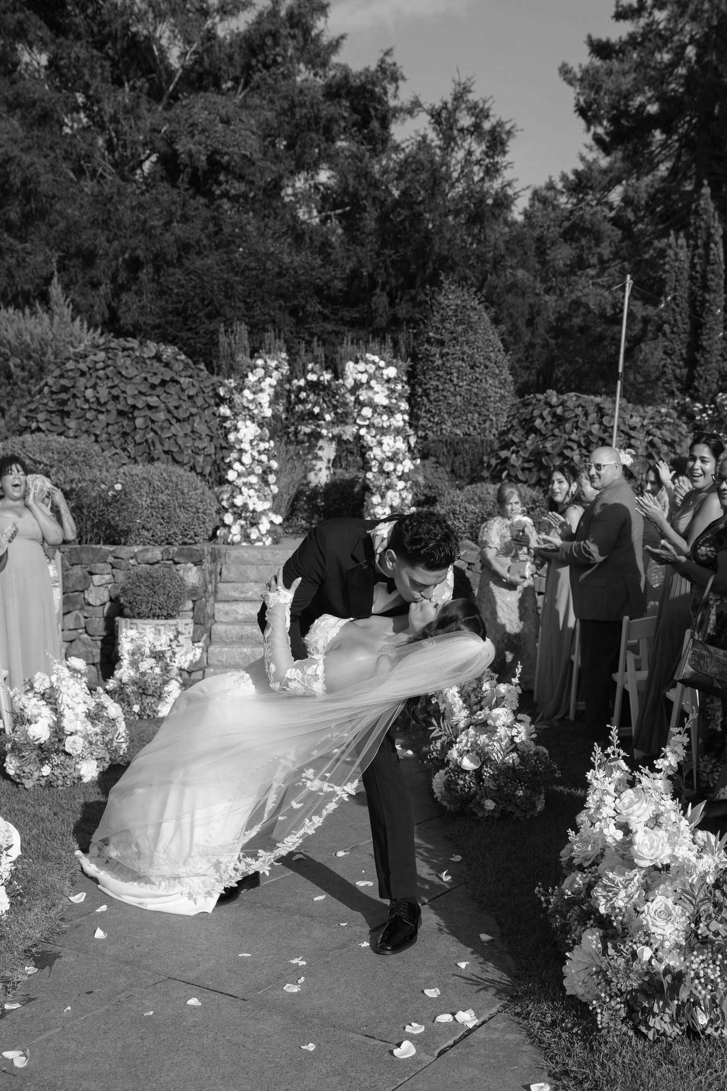 Black and white photo of groom dipping bride during recessional with guests cheering along floral-lined aisle.