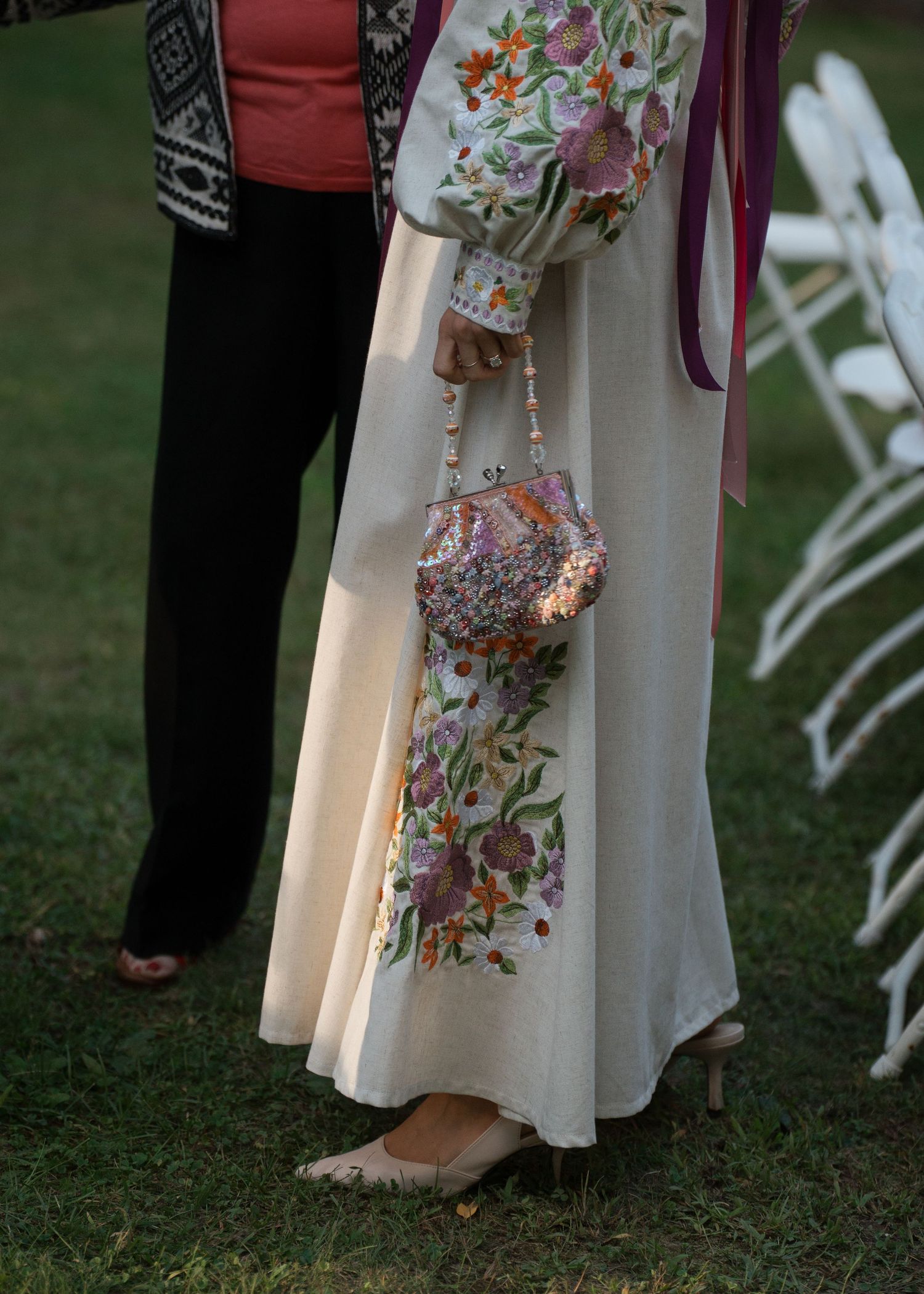 Elegant woman in floral embroidered cream gown holding a beaded vintage purse at an outdoor wedding ceremony.