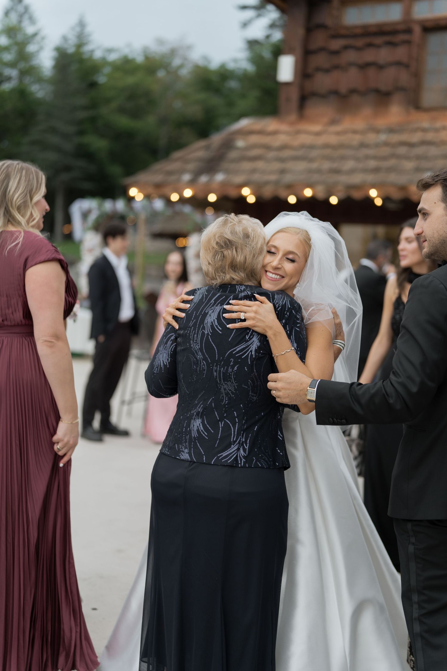 Smiling bride in white veil embracing elderly woman in navy sequin dress at outdoor wedding reception.