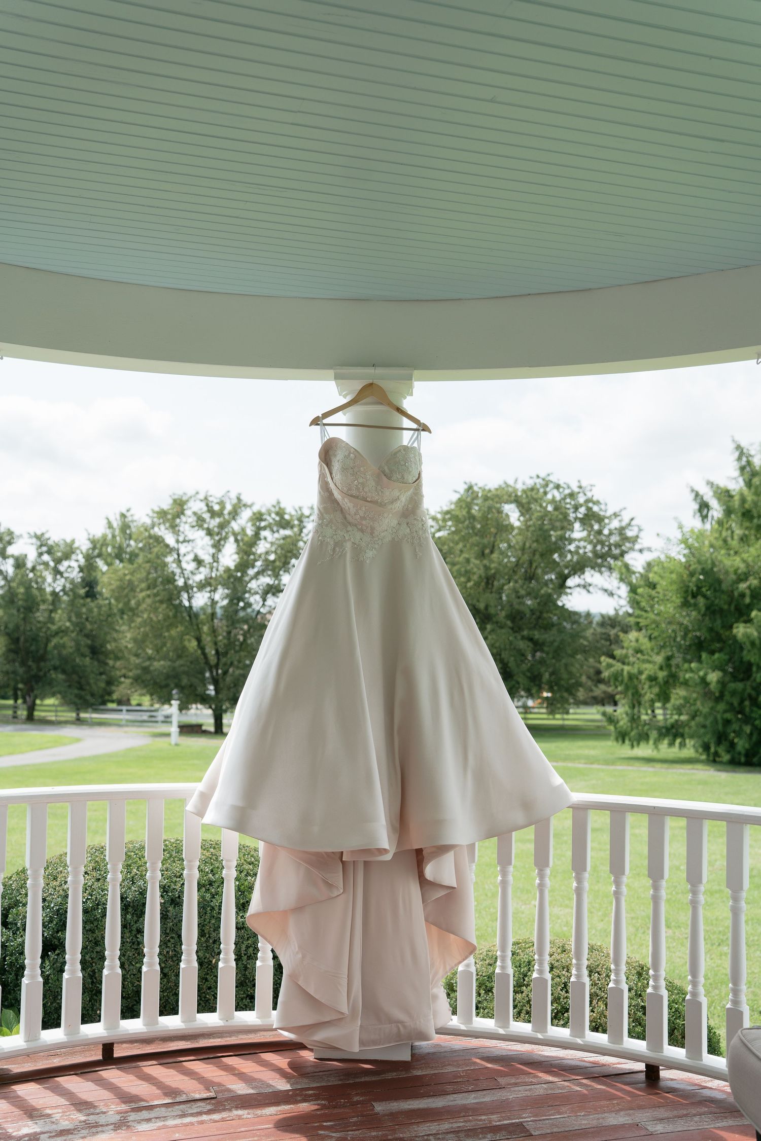 Elegant strapless A-line wedding dress hanging on a wooden hanger in a white gazebo overlooking lush green gardens.