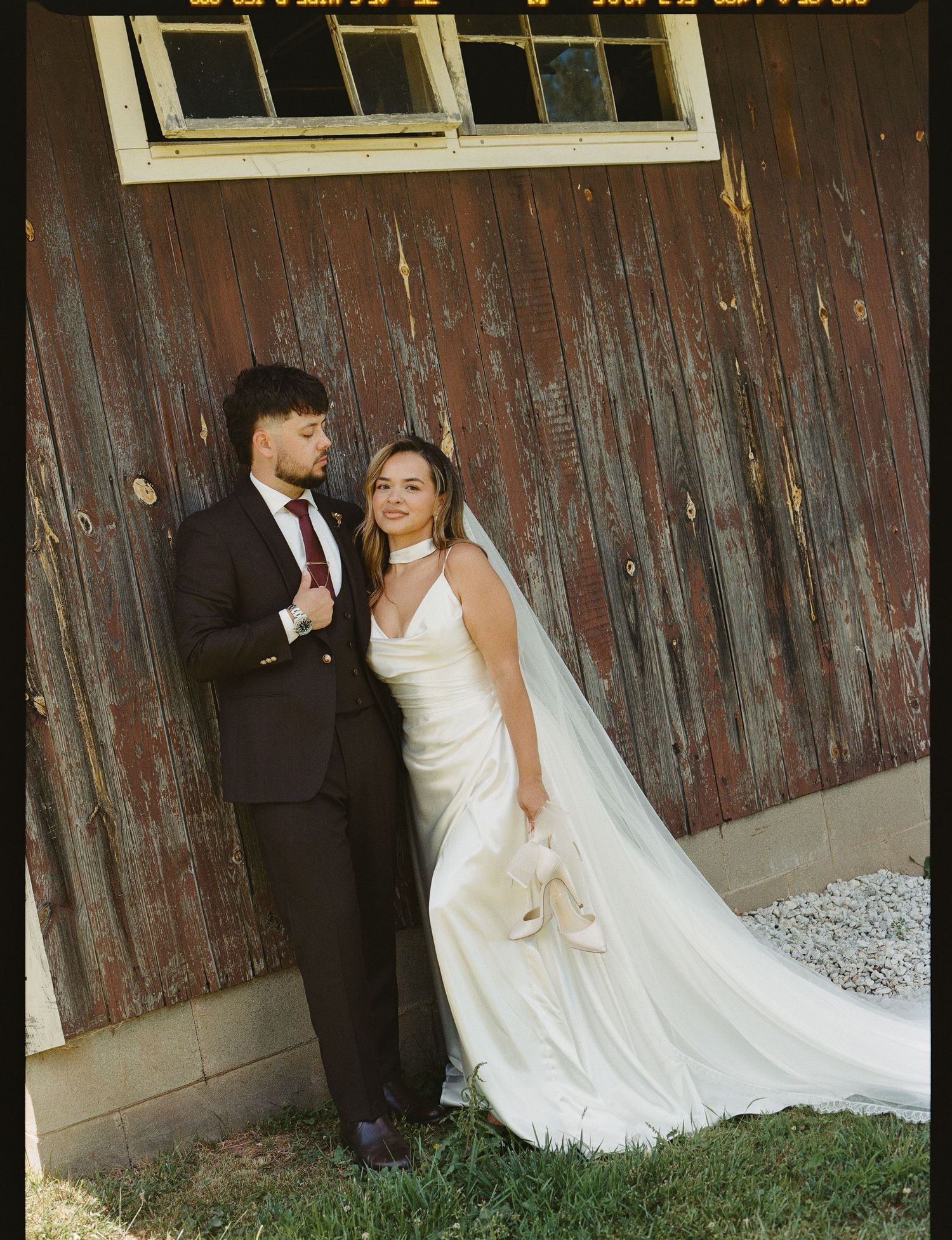 Bride and groom pose together in front of rustic barn, she in white satin gown and veil, he in dark suit.