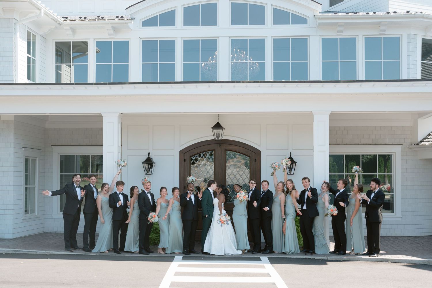 Wedding party poses in front of elegant white venue with large windows and grand entrance doors.