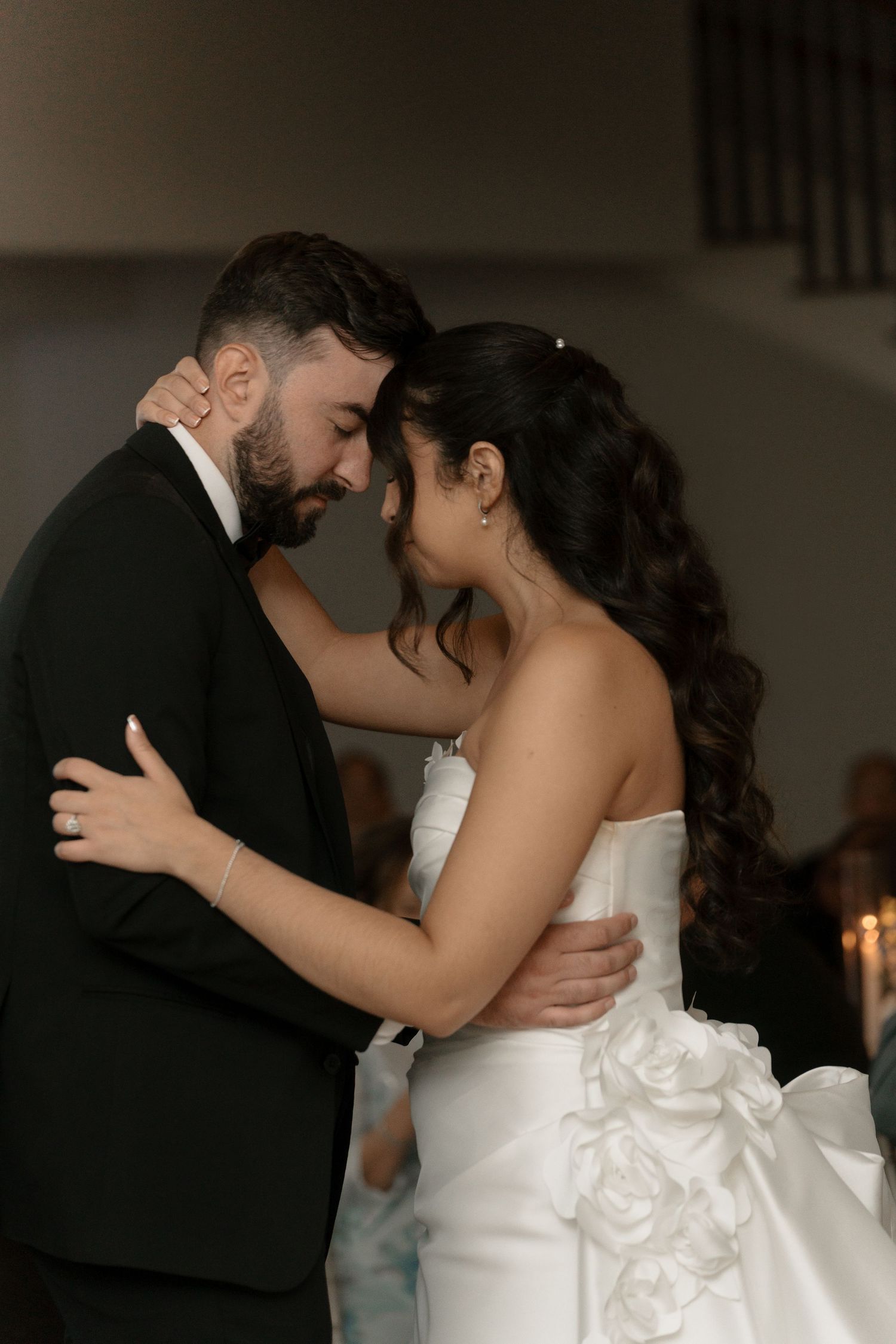 Bride and groom share an intimate first dance moment, foreheads touching, in a softly lit wedding venue.