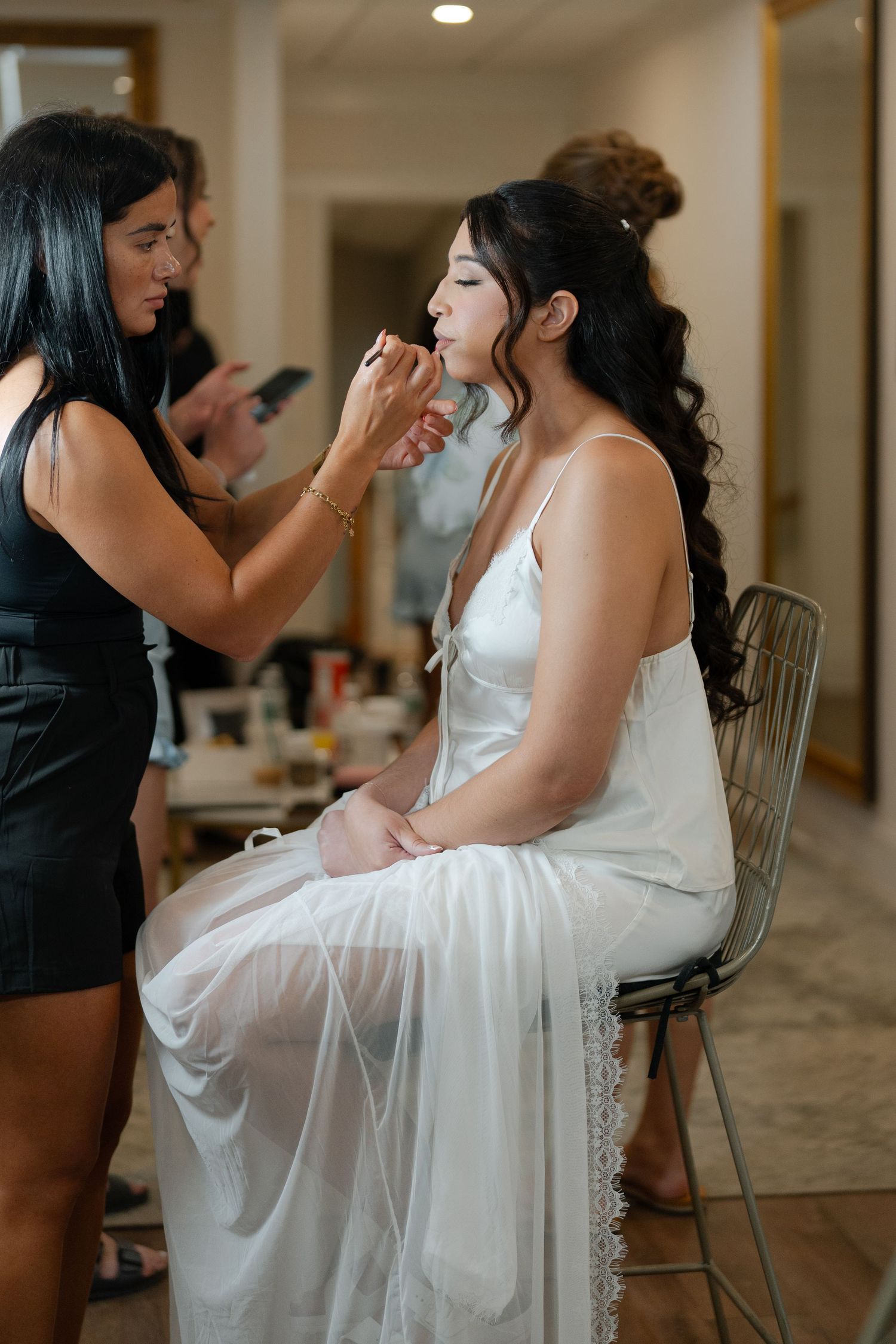 Makeup artist applying lipstick to bride in white dress seated in chair during wedding preparations.