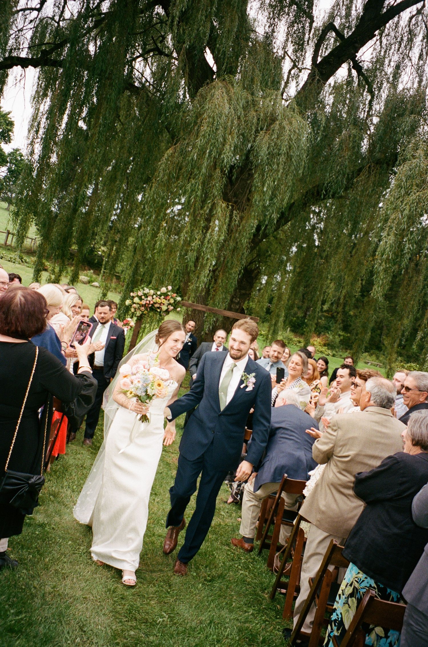 Wedding guests gather under a weeping willow tree during an outdoor celebration ceremony.