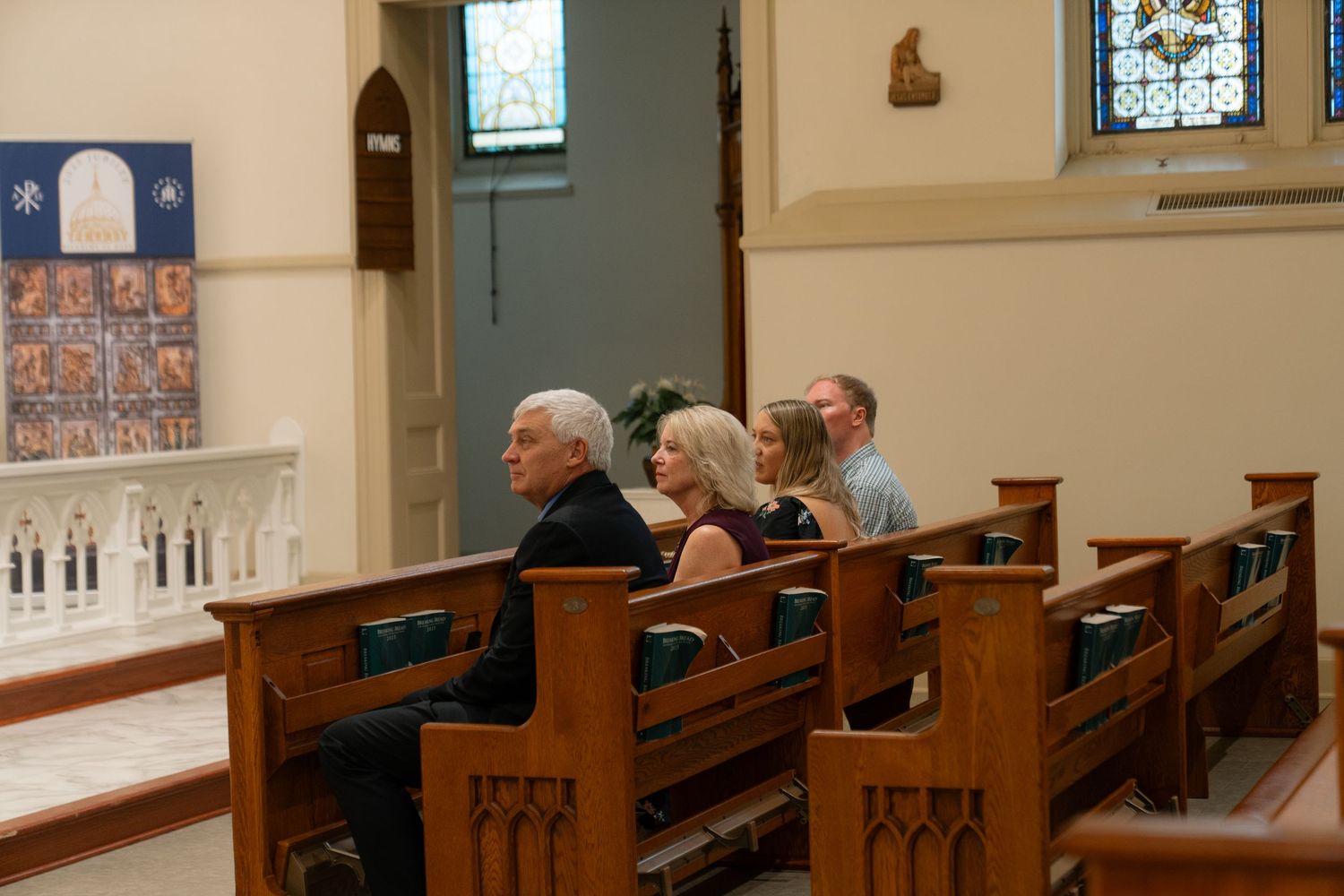 People sit quietly in wooden pews inside a traditional church with stained glass windows and white walls.