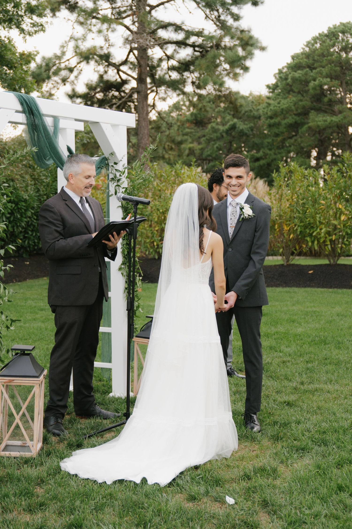 An outdoor wedding ceremony takes place on a grassy lawn with pine trees and lantern decorations at sunset.