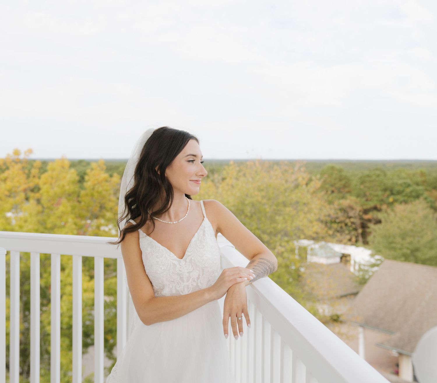 A bride in a white wedding dress stands on a white balcony overlooking a scenic autumn landscape.